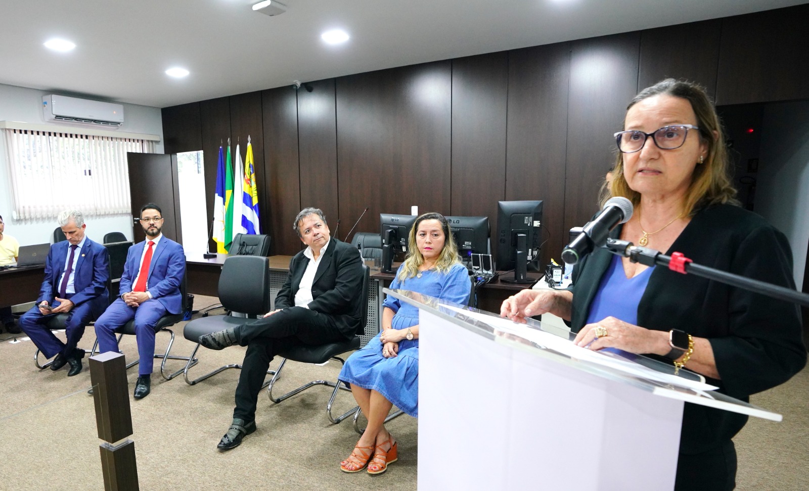Desembargadora Maysa Vendramini Rosal discursa na tribuna. Ela usa um blazer preto e uma blusa azul observada por três homens e uma mulher