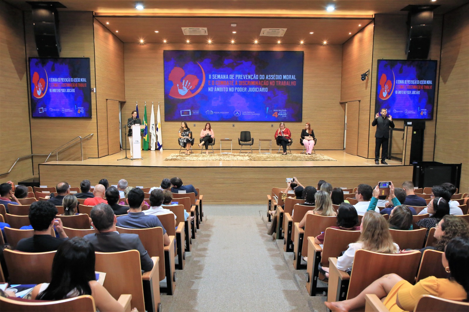 Imagem colorida de pessoas sentadas em cadeiras num auditório com palestrantes à frente um telão colorido