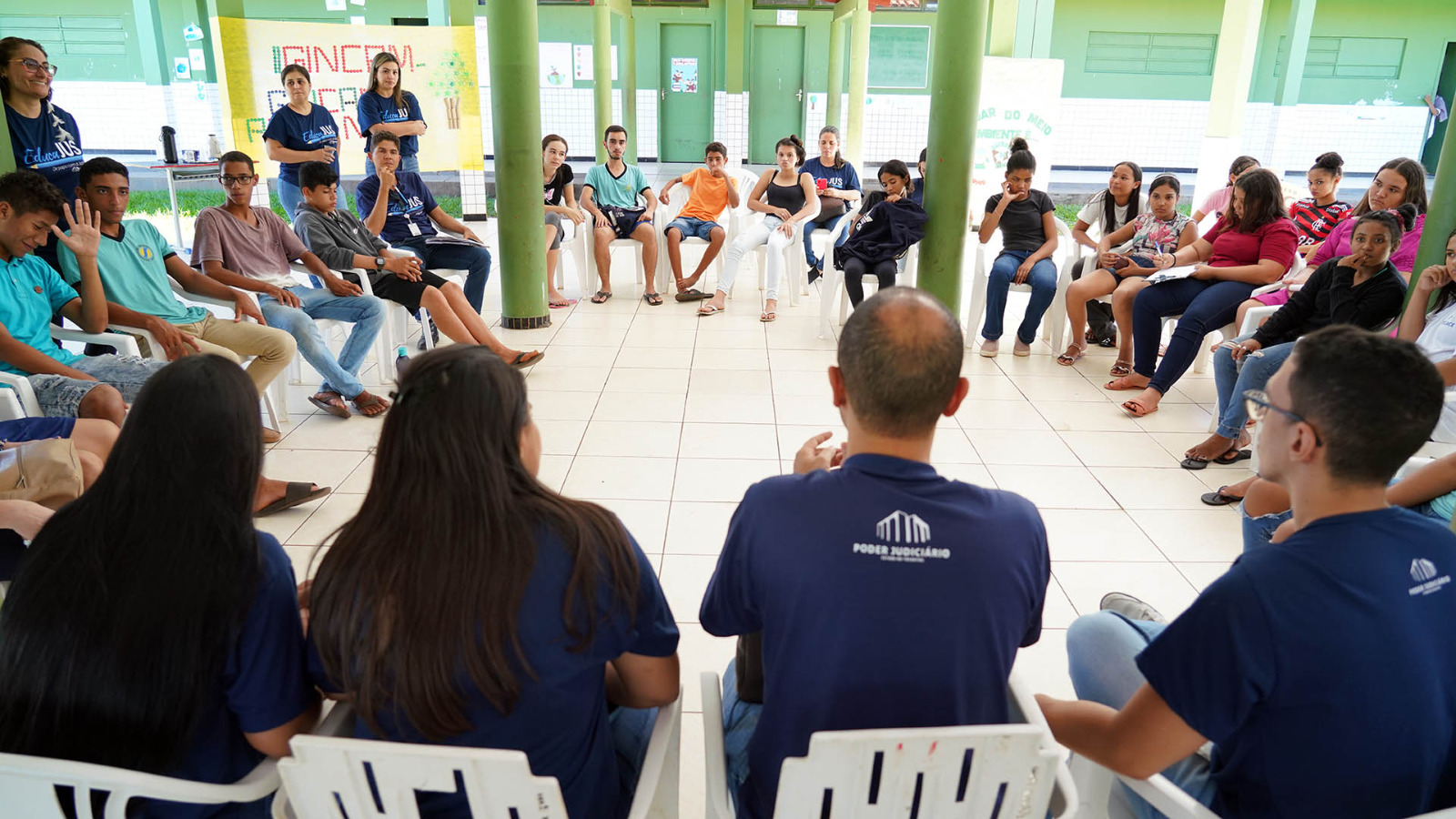 Juíza em pé com mais duas pessoas à frente de outras pessoas sentadas em círculo em uma escola durante discussão sobre cidadania com alunos