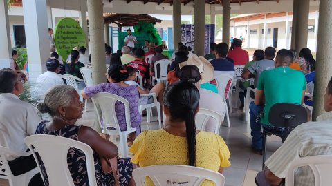 Imagem mostra várias pessoas sentadas em cadeiras de plástico branca e ao fundo uma pessoa fala no microfone