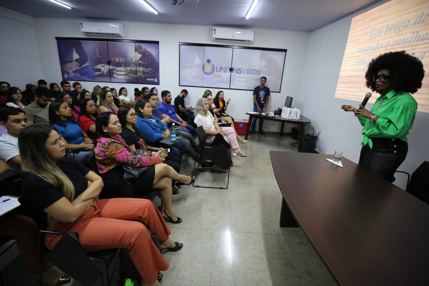 Imagem colorida de mulher negra vestindo verde falando para um auditório lotado de estudantes da Unitins 
