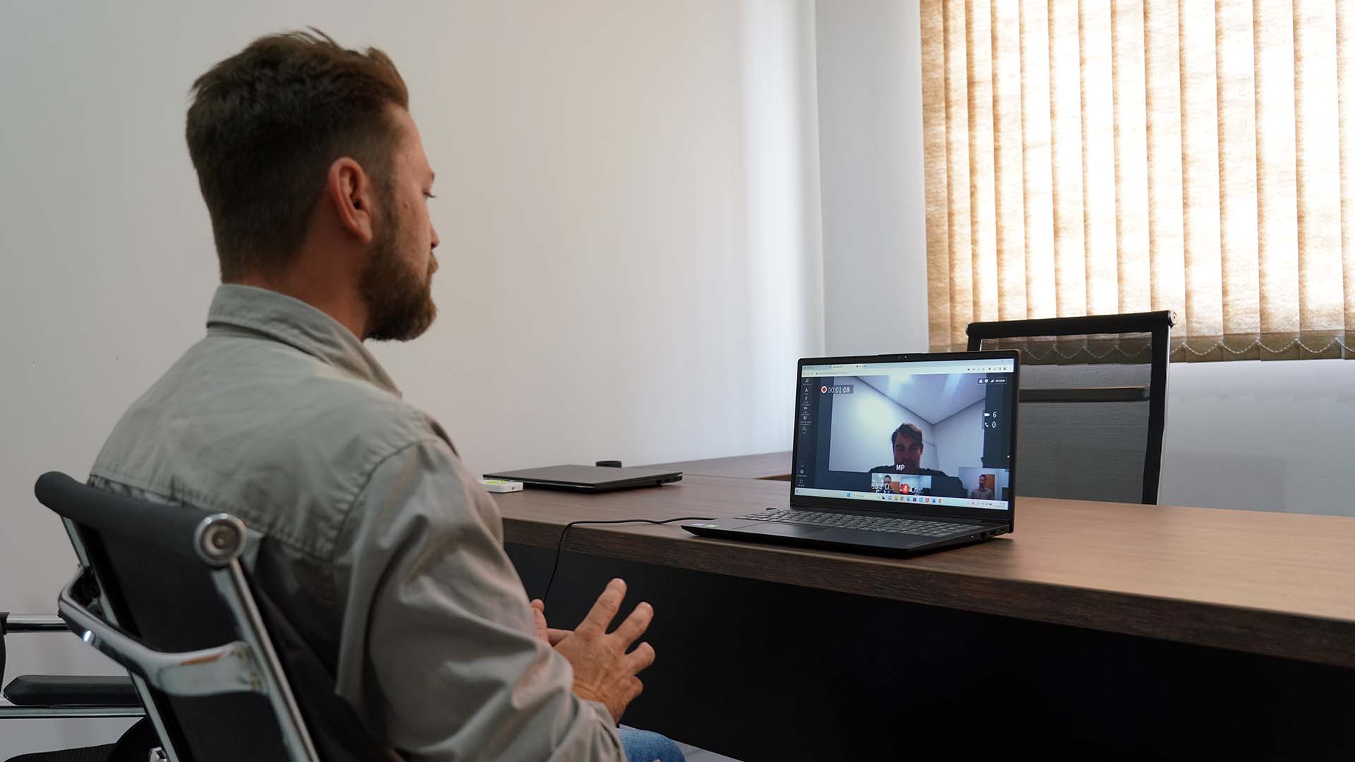 Homem branco de camisa e barba sentado em frente a um notebook conversa com juiz durante audiência