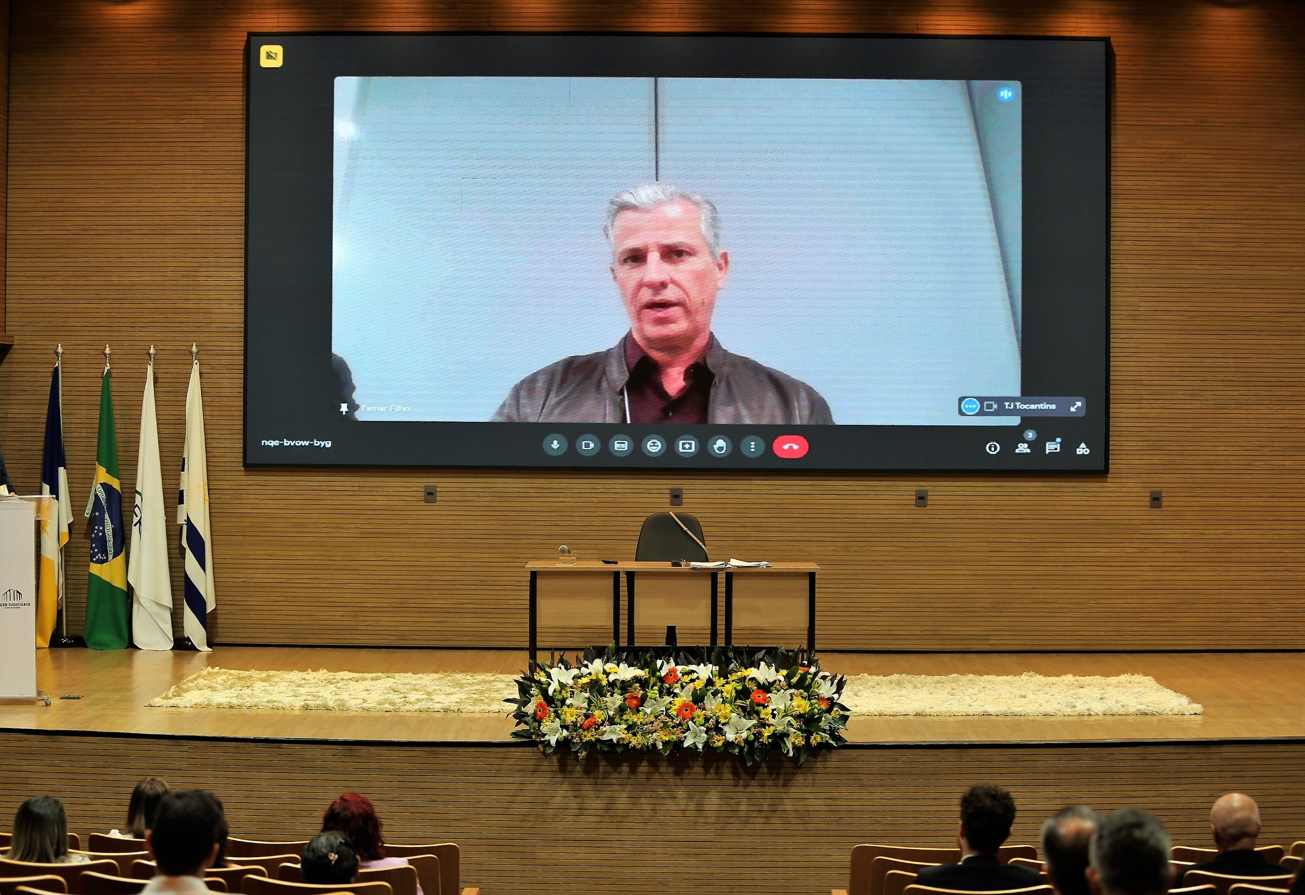Fotografia colorida mostra um homem branco e grisalho em destaque num telão dentro de um auditório 