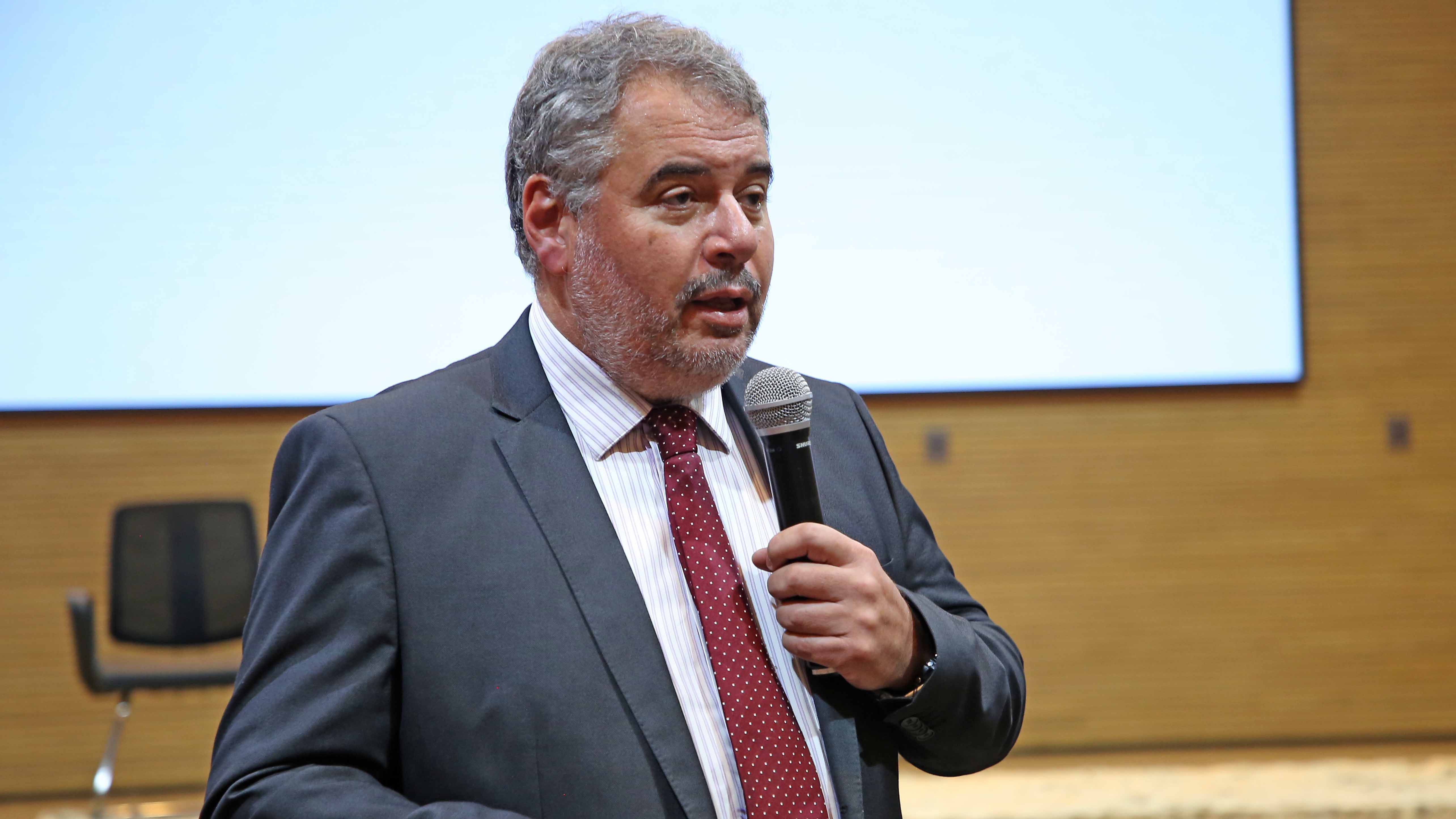 Camal Godoy usa terno e gravata e segura um microfone