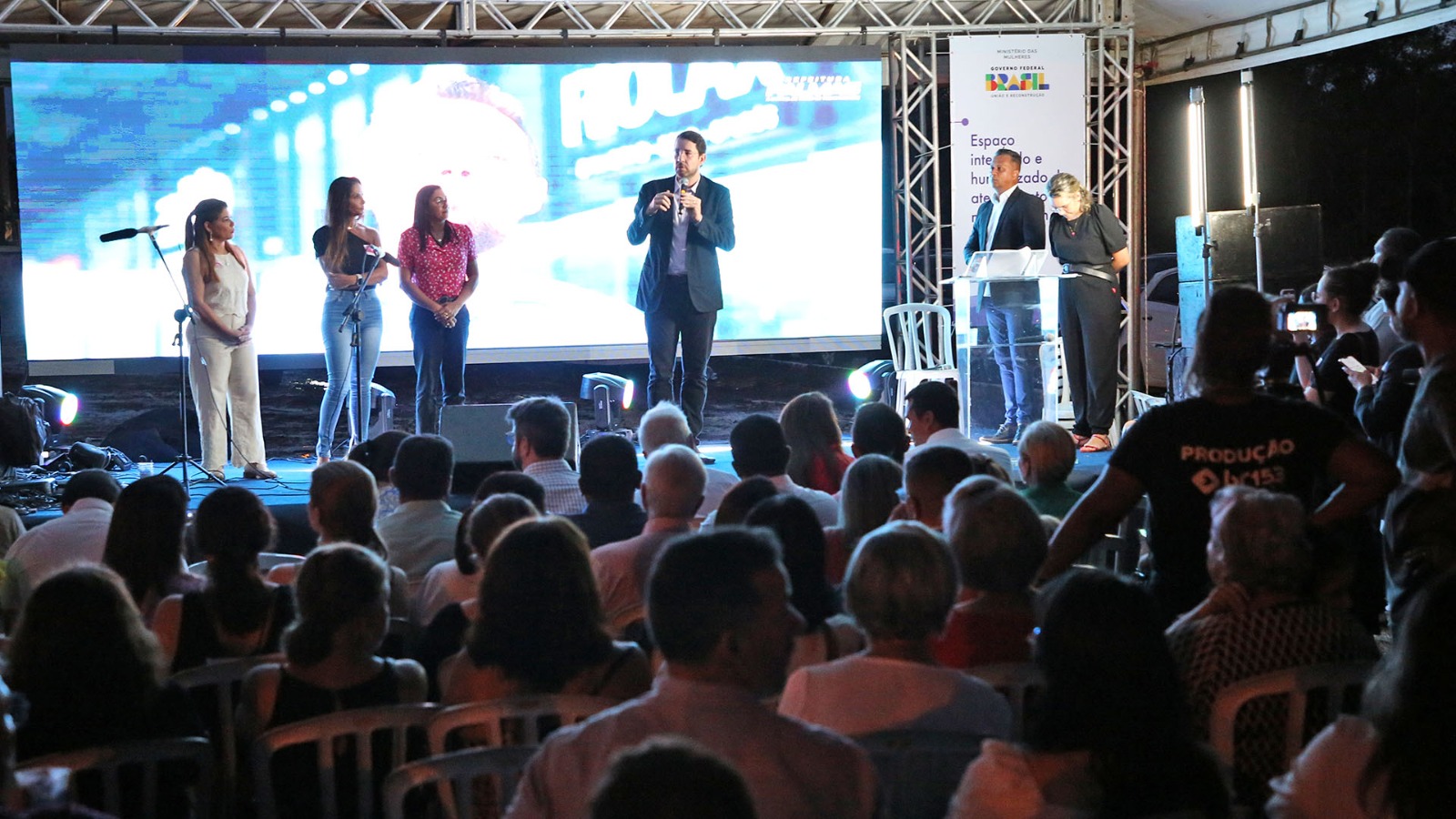 Imagem colorida que mostra um grande público, em espaço aberto, vendo autoridades no palco