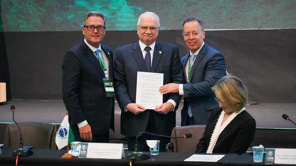 Imagem mostra três homens em pé, segurando um papel, e ao lado uma mulher sentada olhando para eles