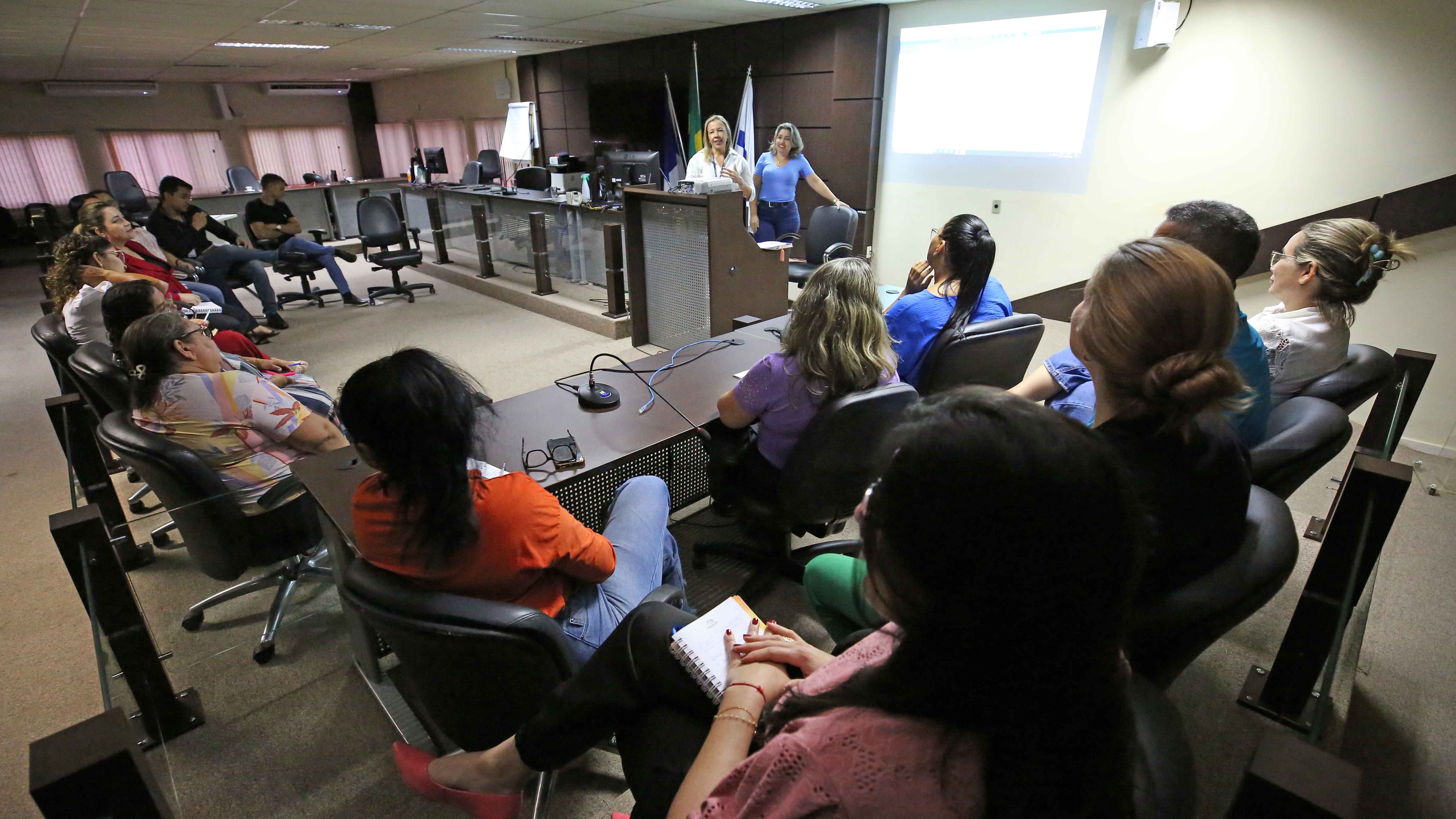 Homens e mulheres sentados durante treinamento ministrados por duas mulheres que estão em pé, uma de camisa branca e cabelos loiros soltos e outra de roupa azul e cabelos castanhos soltos, ambas atrás de uma mesa alta