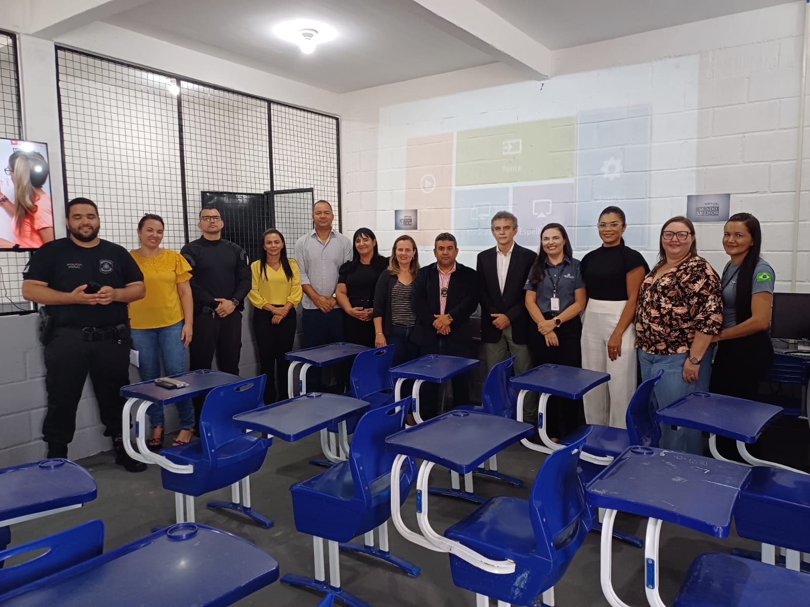 Imagem mostra carteiras escolares com cor azul à frente e atrás pessoas em pé posando para foto