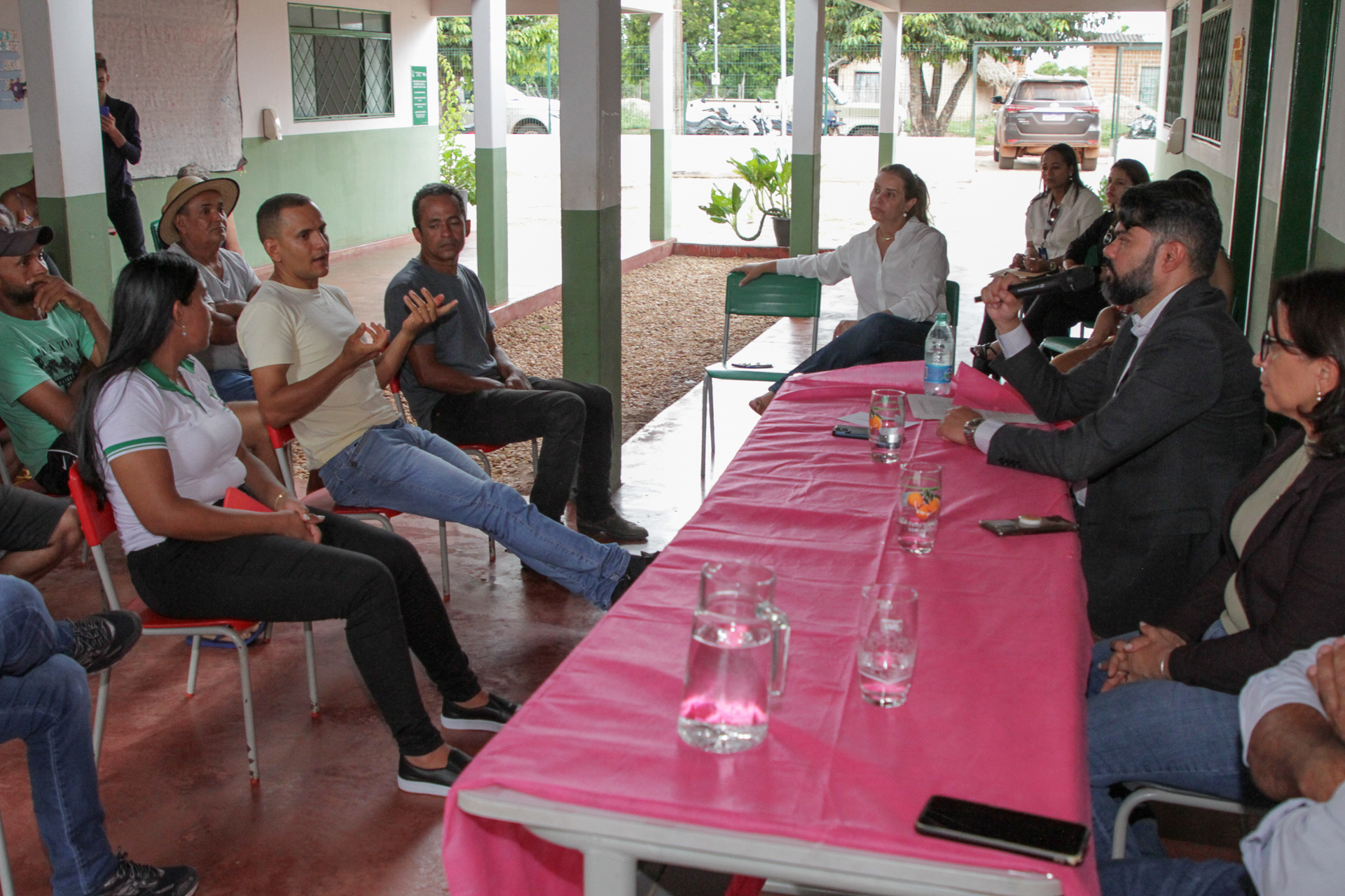 Fotografia colorida de pessoas sentadas dos dois lados de uma longa mesa retangular, de um lado pessoas do poder judiciário e do outro a população do povoado, enquanto um homem conversa com outro que está do outro lado da mesa, em sua frente, sobre a regularização fundiária  