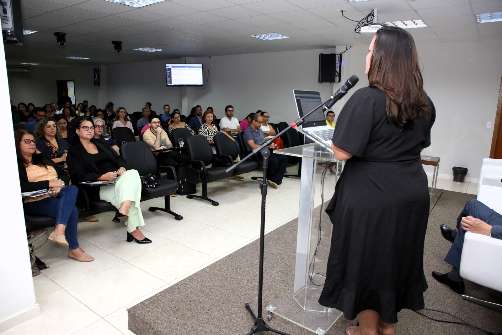 Fotografia colorida de uma mulher em pé em um palanque, usando vestido preto e com cabelo, ministrando a aula de Automatização de Localizadores no Sistema Eproc, enquanto os ministrados estão sentados na plateia assistindo a aula