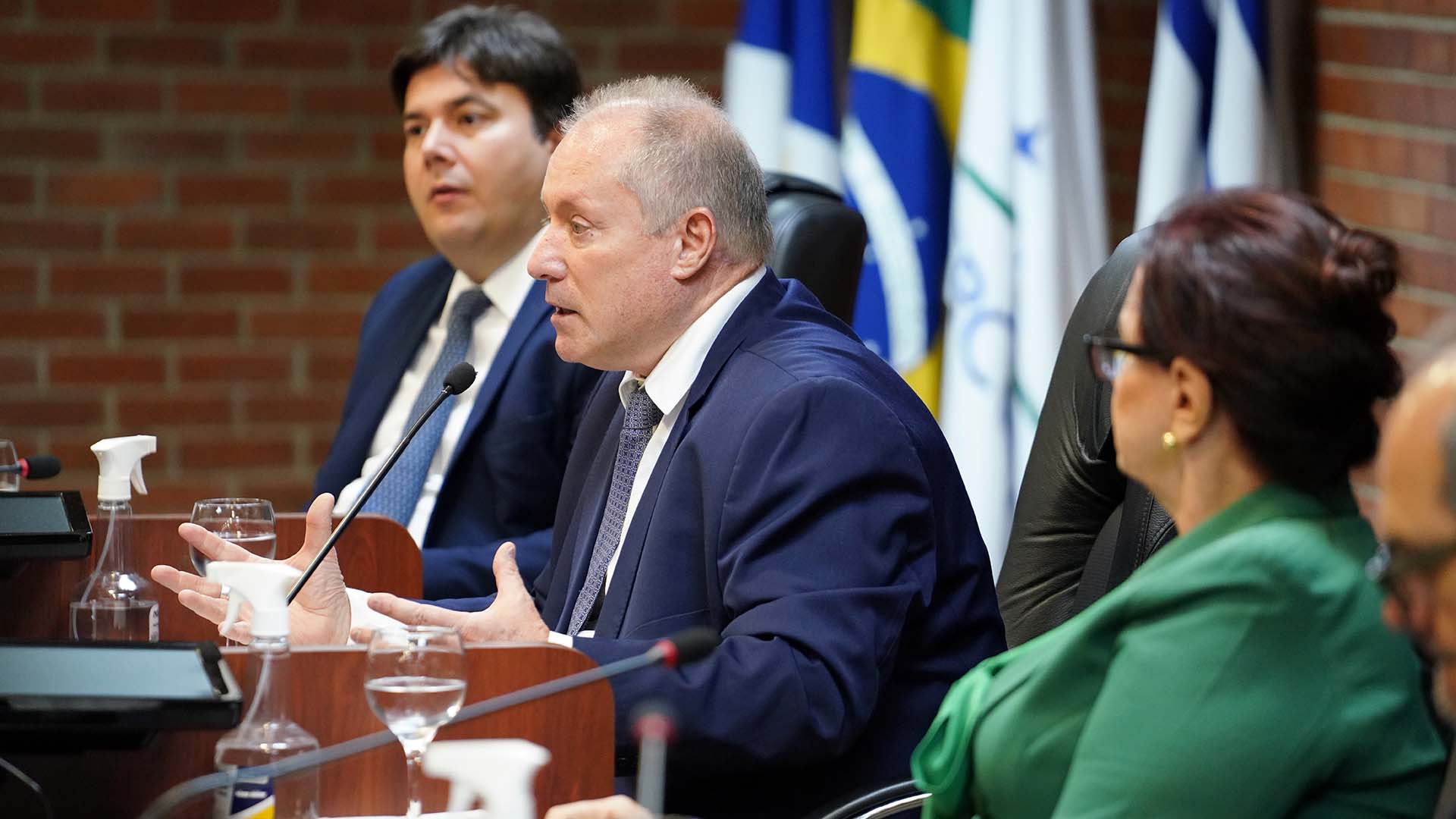 Imagem mostram três pessoas brancas,  sendo uma mulher; todas estão sentadas; um fala e dois observam