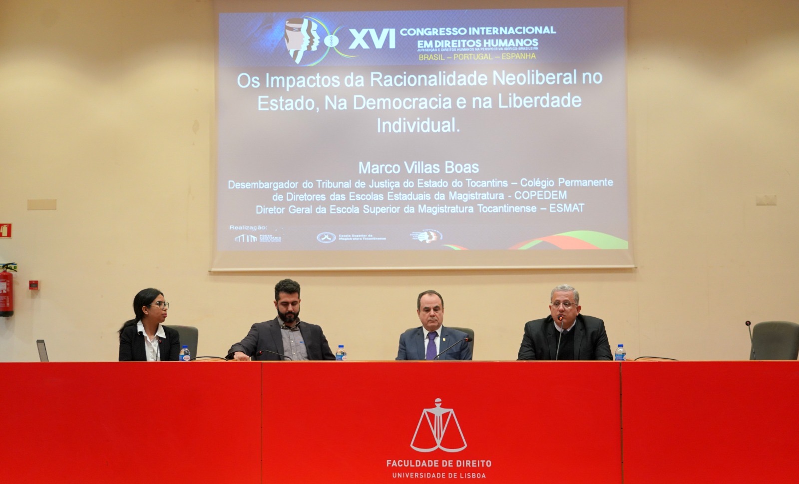 Fotografia colorida de quatro pessoas sentadas atrás de uma mesa no XVI Congresso Internacional de Direitos Humanos, com um holograma repleto de informações do evento passando ao fundo delas 