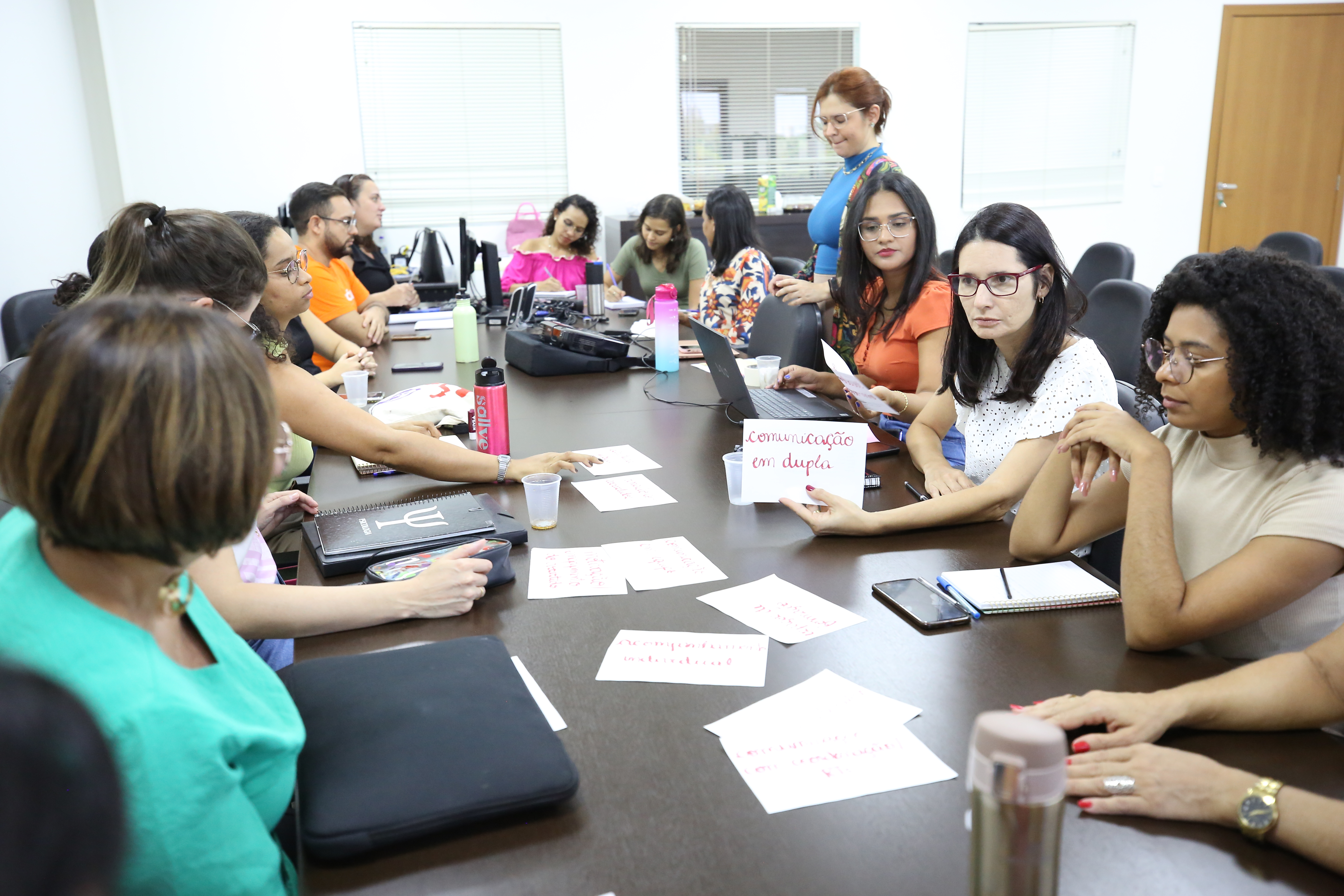 Numa sala, várias mulheres, profissionais que integram a equipe multiprofissional do projeto, reunidas em volta de uma mesa