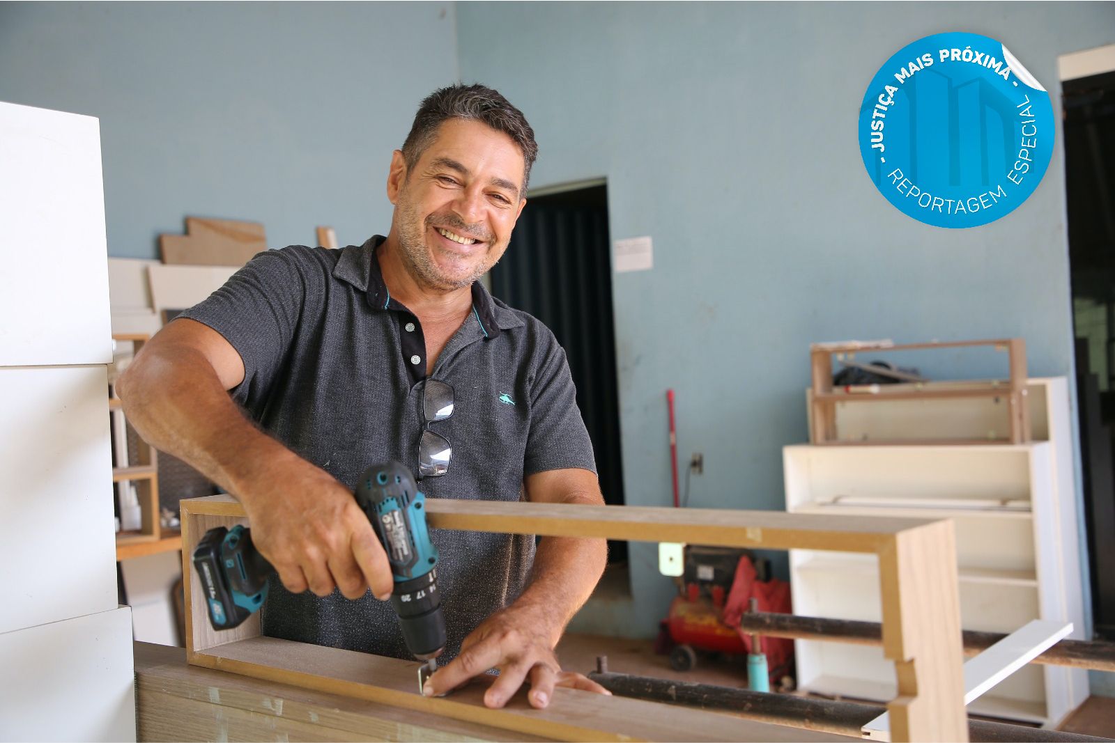 José Humberto, trabalhando em sua oficina de marcenaria; ele porta um equepamento de trabalho, está sorrindo e usa camisa cinza