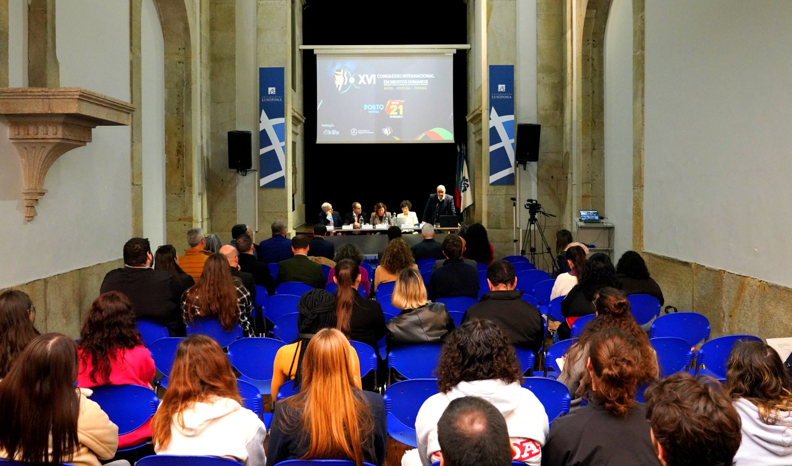 Fotografia colorida onde pessoas estão sentadas em cadeiras azuis assistindo o XVI Congresso Internacional dos Direitos Humanos 