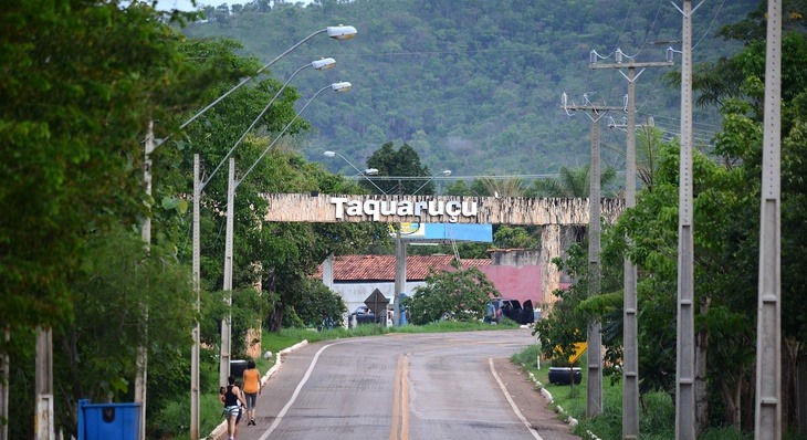 Fotografia colorida do portal da cidade de Taquaruçu, com muitas arvores em volta da estrada que segue para sua entrada, com uma casa de fundo, e a serra da cidade atrás  