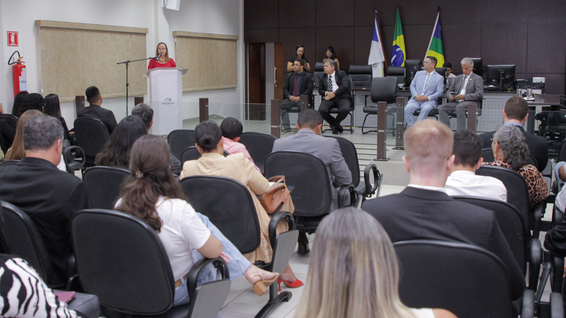 Fotografia colorida do público assistindo a desembargadora Maysa Rosal discursando sobre um palanque de madeira escura , usando uma camisa vermelha na Correição Geral Ordinária de Augustinópolis 