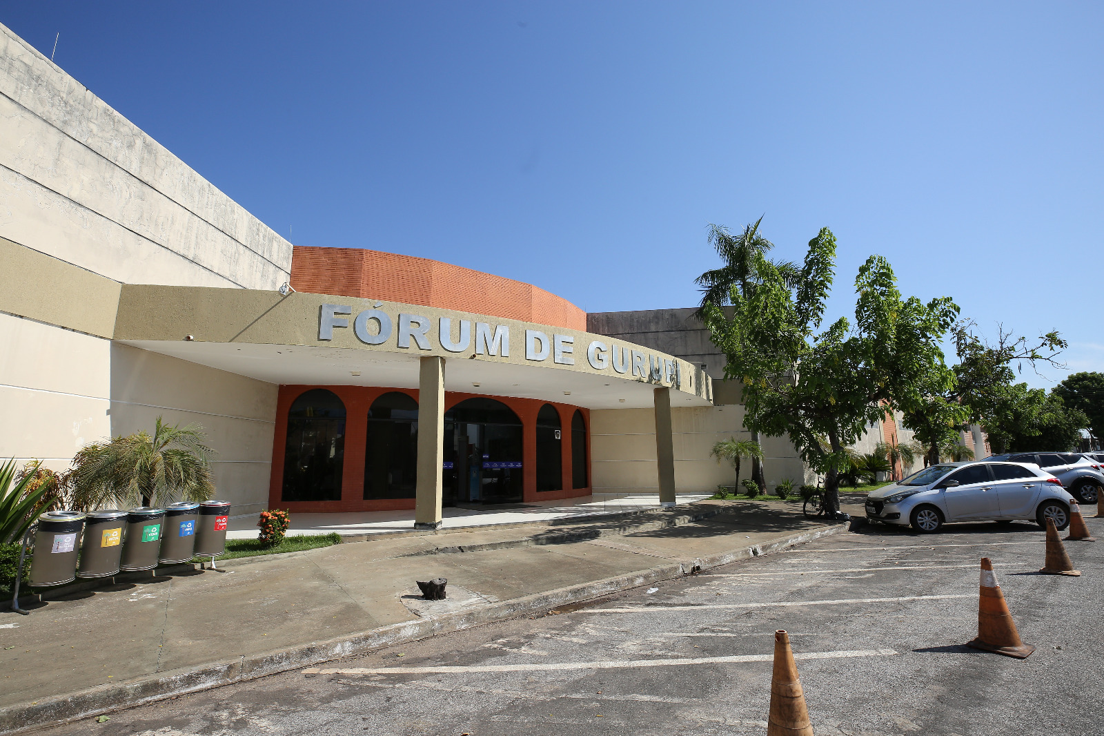 Fachada do Fórum da Comarca de Gurupi; paredes pintadas nas cores bege e vermelha; à frente, parte do estacionamento e lixeiras da coleta seletiva