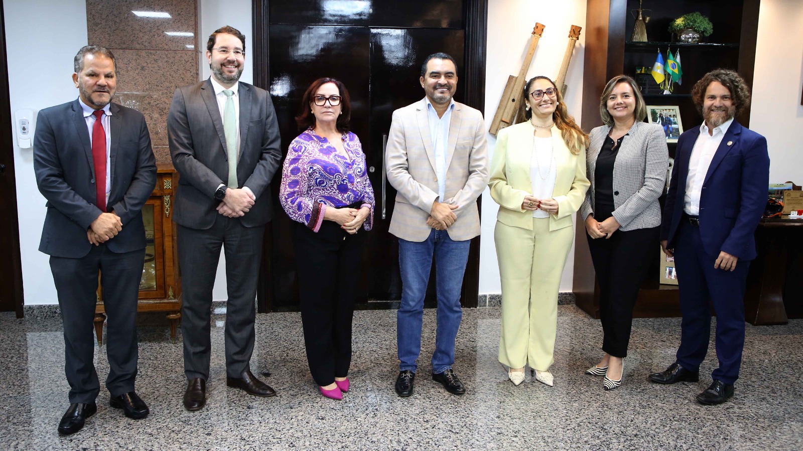 Foto posada com sete pessoas em pé,  sendo três mulheres e quatro homens; eles estão em uma sala ampla que é o gabinete do governador