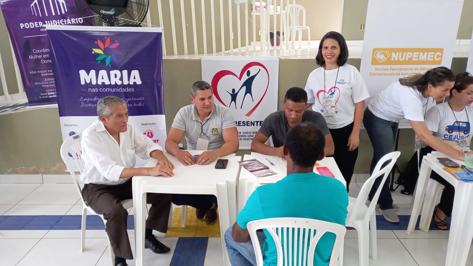 uma mulher de pé posa para foto junto a uma mesa branca onde estão sentados quatro homens
