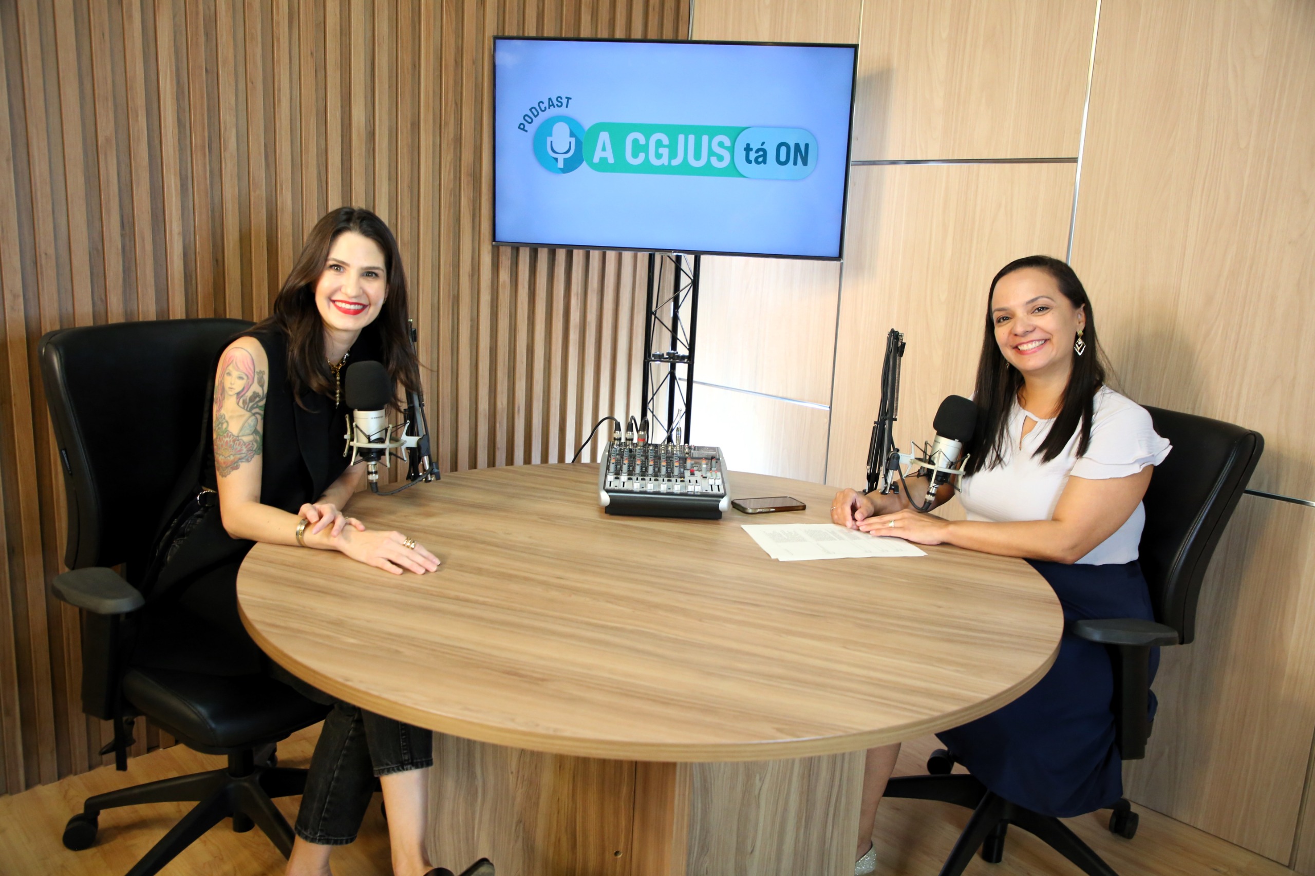Fotografia colorida onde duas mulheres estão sentadas uma de frente para a outra em uma mesa redonda de madeira clara, com uma televisão ligada de fundo e uma parede também de madeira clara 