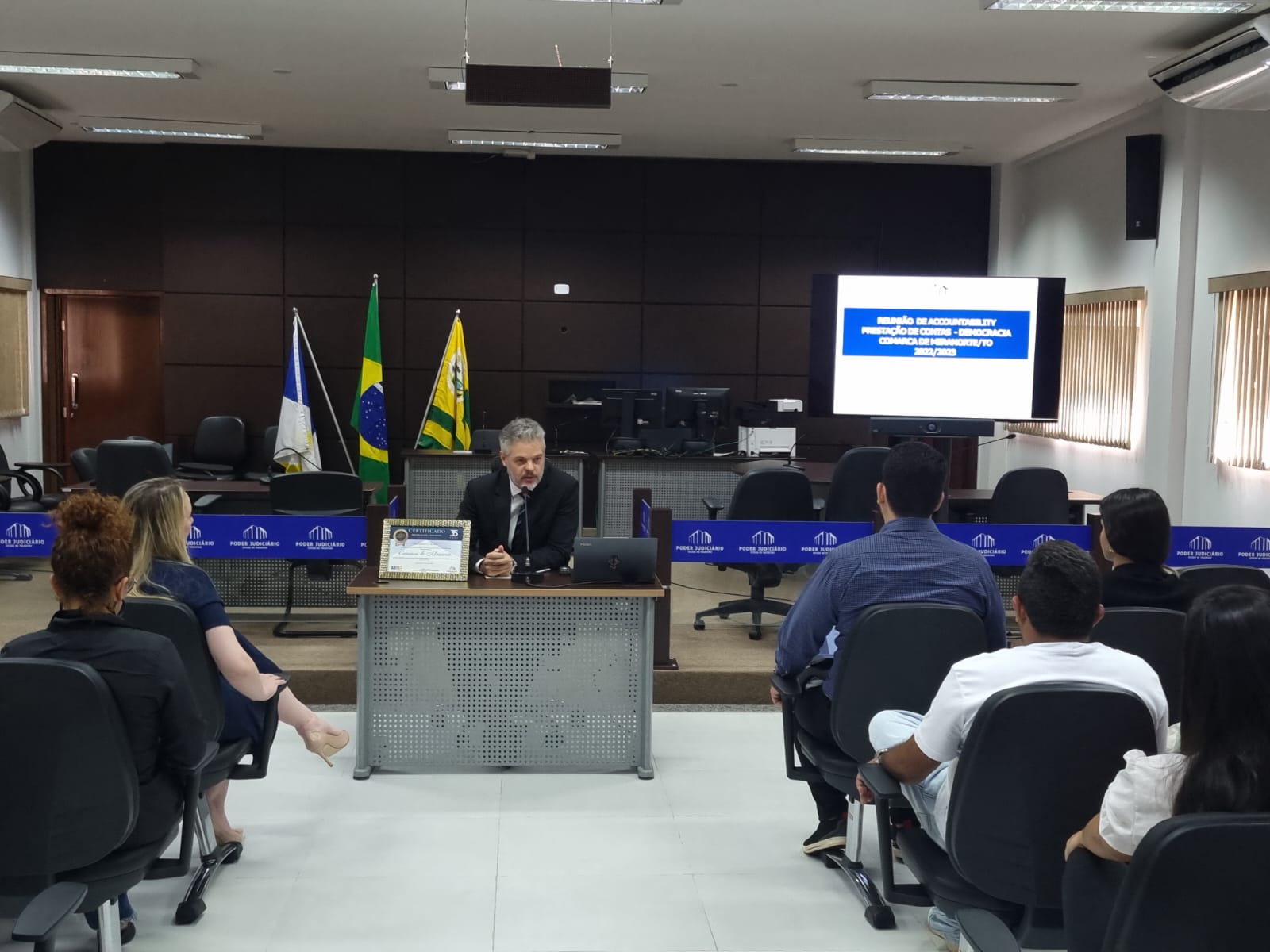 juiz Ricardo Gagliardi, sentado à frente, ele tem cabelos e barba grisalhos  e usa terno escuro e camisa branca; à sua frente várias pessoas sentadas no auditório