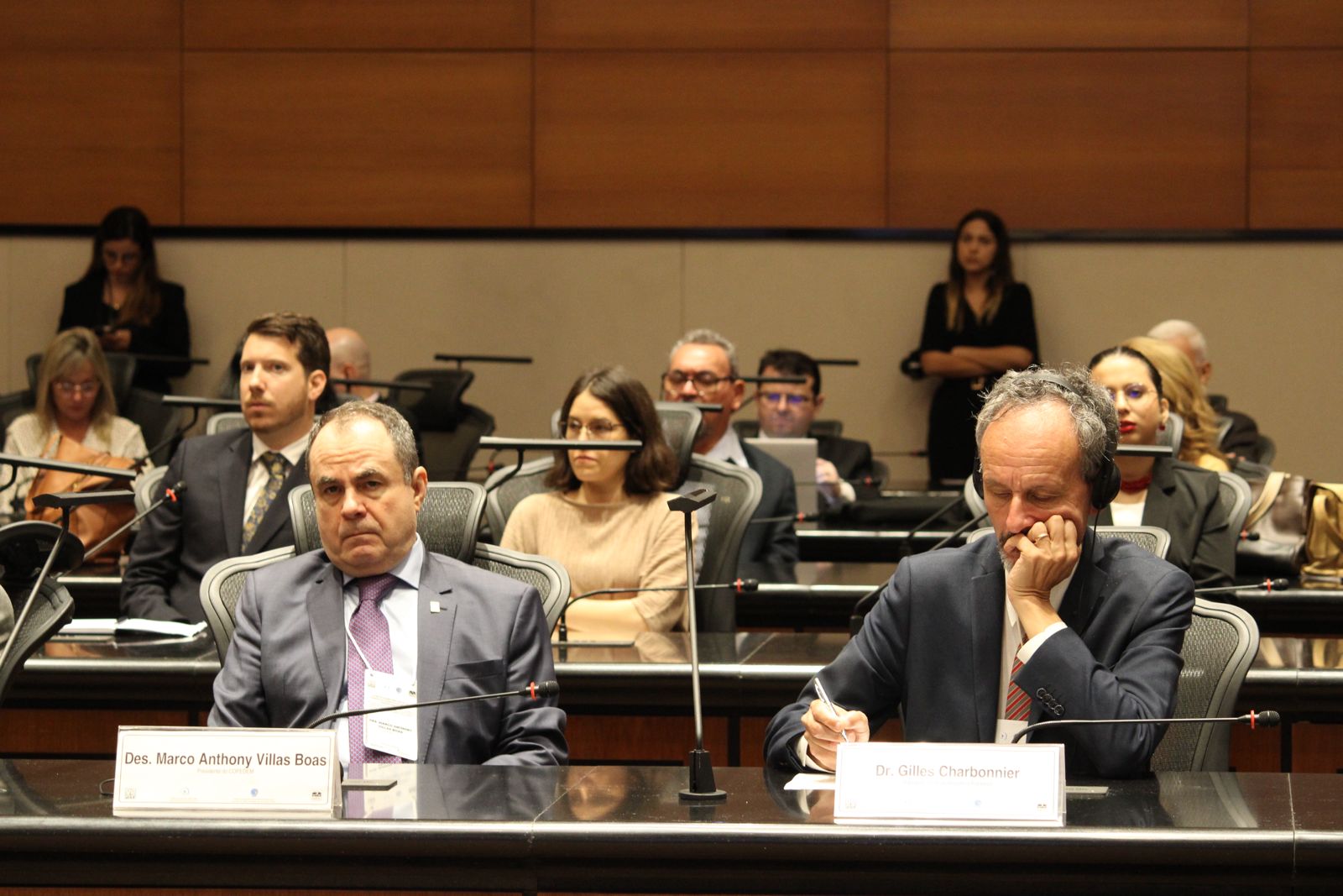 Imagem mostra à frente dois homens sentados em mesas identificadas em atrás várias outras pessoas