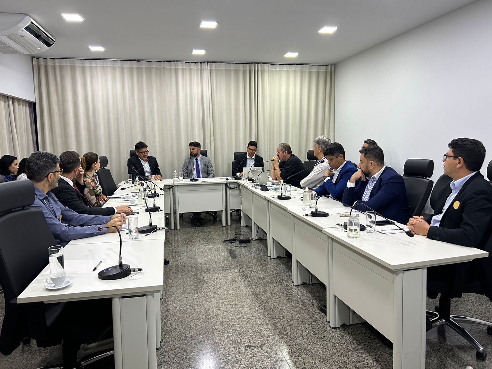 Imagem mostra uma sala de reunião, uma mesa em formato de U e várias pessoas sentadas; à frente de cada uma existe um microfone