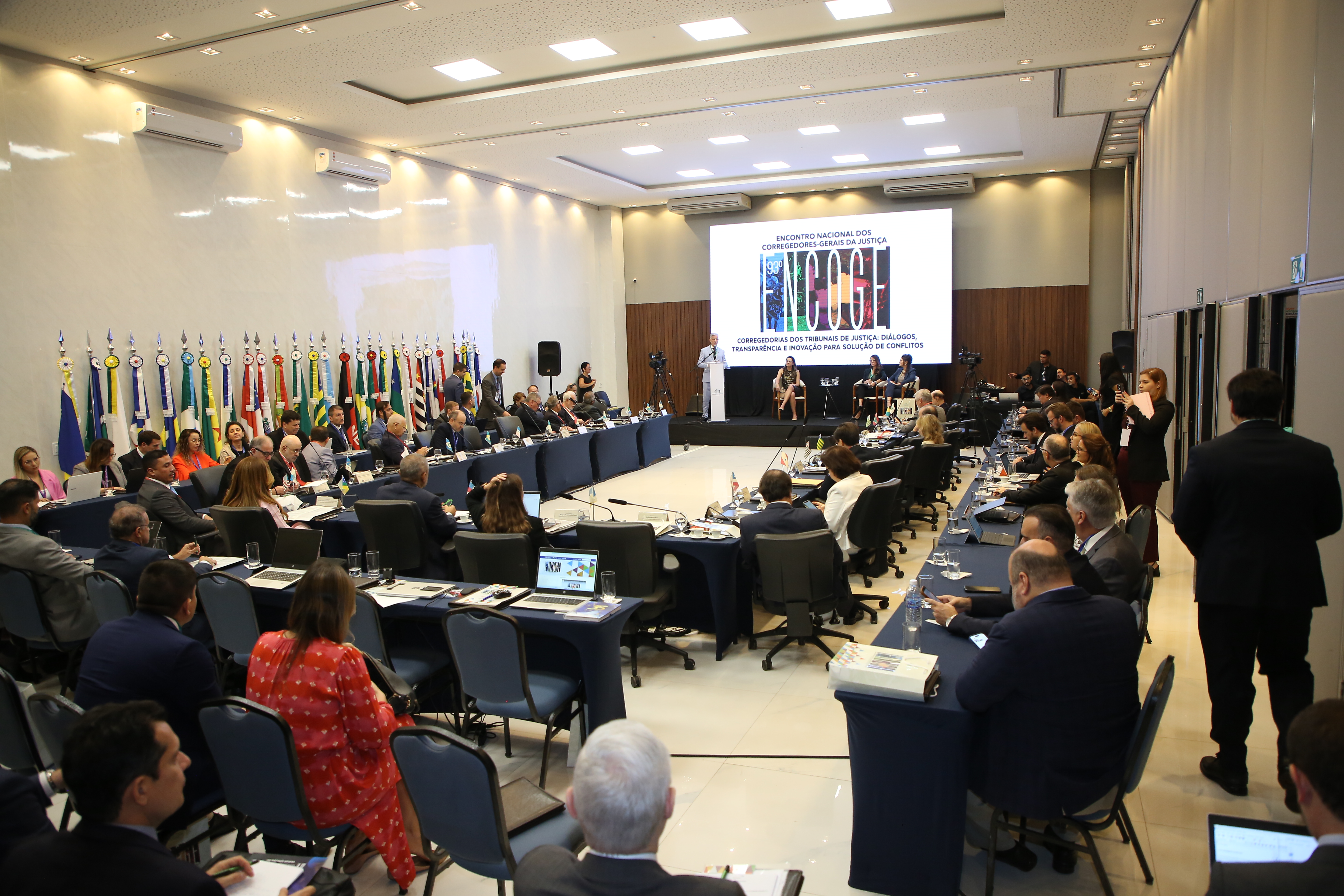 Fotografia colorida onde várias pessoas estão sentadas em cadeiras, em um entorno no formato retangular, olhando para um painel escrito ENCOGE