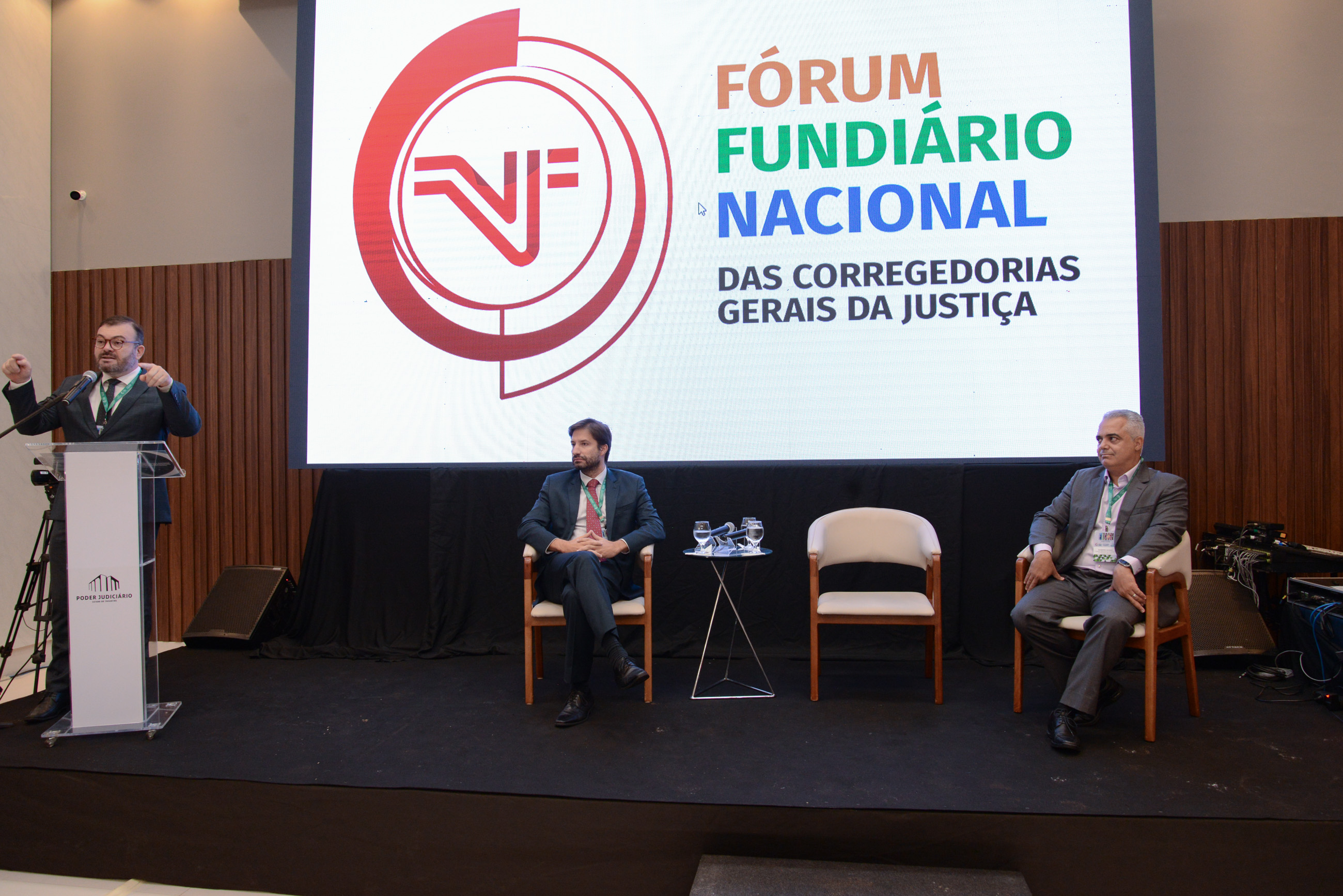 Imagem colorida de dois homens sentados em uma cadeira com um painel ao fundo escrito Fórum Fundiário Nacional e um outro homem de barba falando em um púlpito