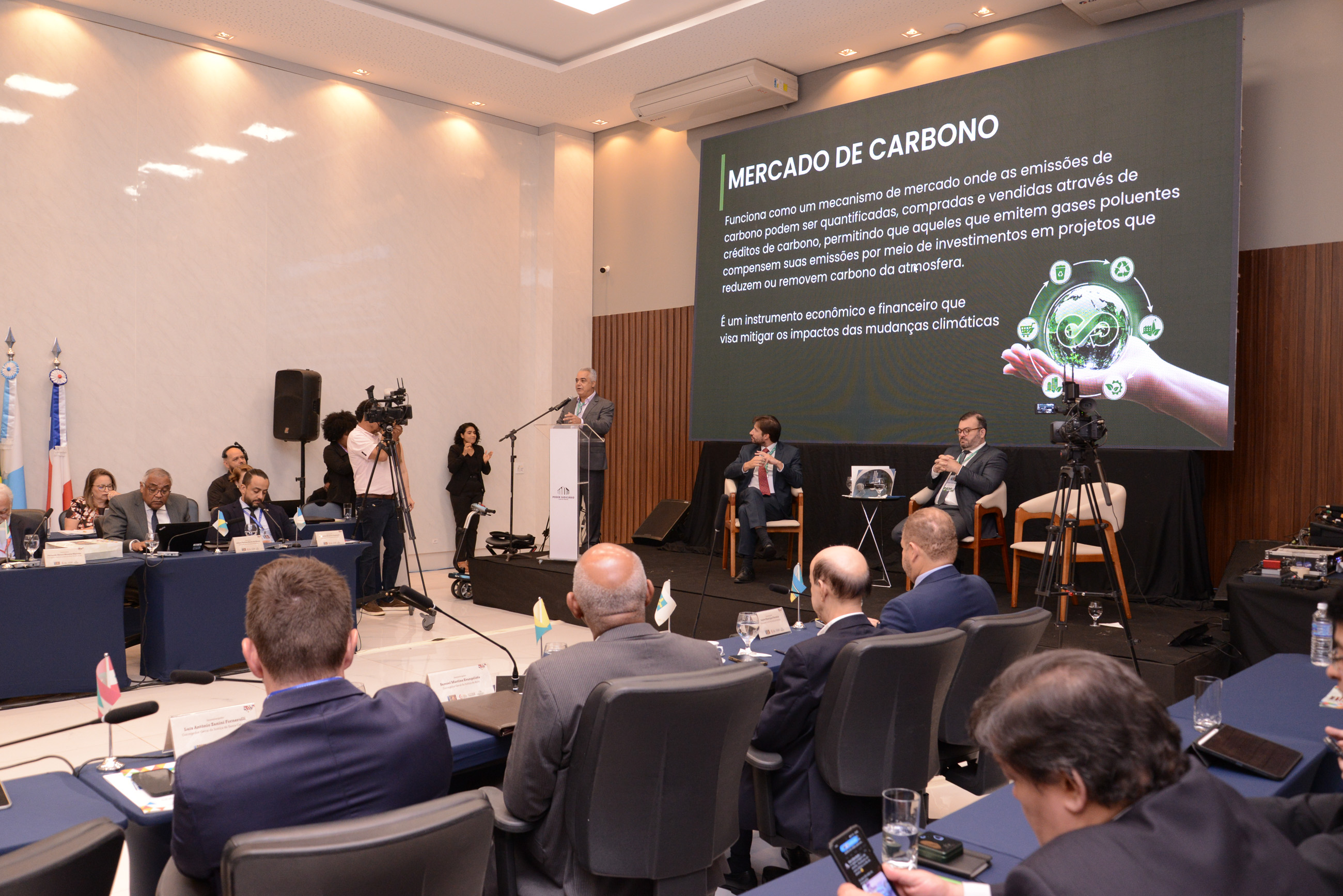 Imagem colorida e um salão com autoridades sentadas à mesa e ao fundo um homem moreno fala em um microfone com um telão no qual lê-se Mercado de Carbono 