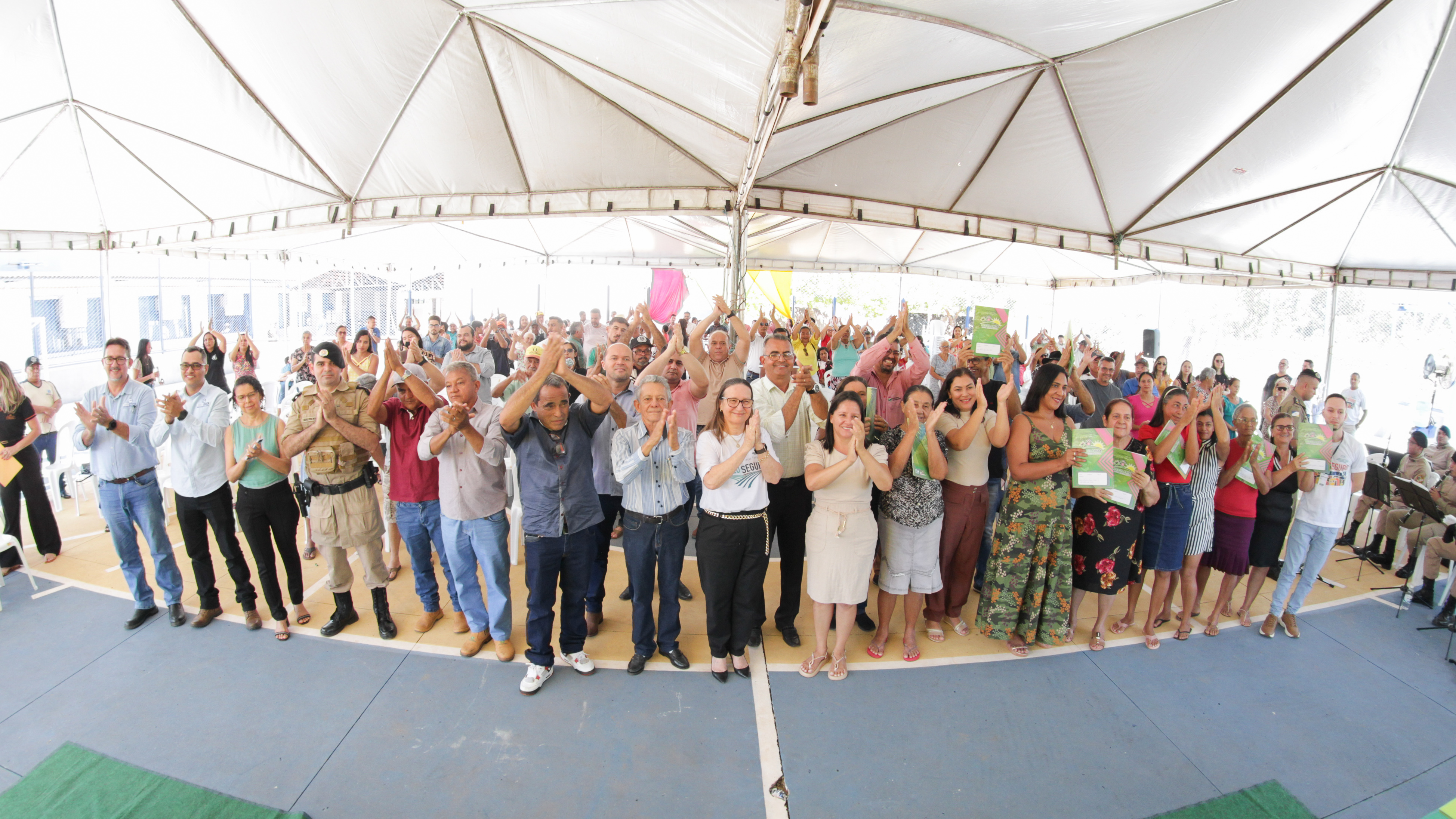 Fotografia colorida onde um grande grupo de pessoas estão reunidos em baixo de uma grande lona branca aplaudindo