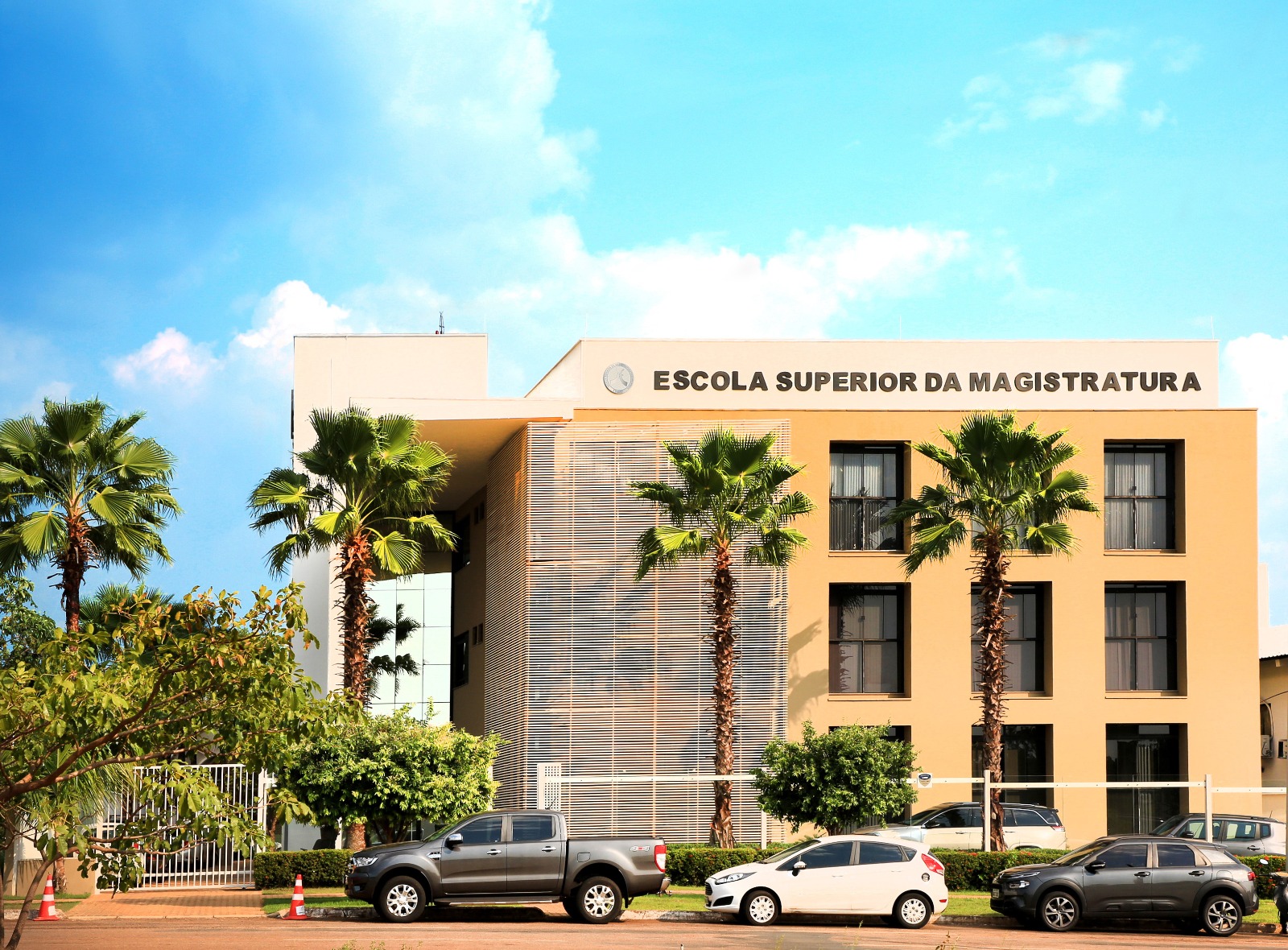 Fotografia colorida da frente da Esmat, com palmeiras e carros na frente 