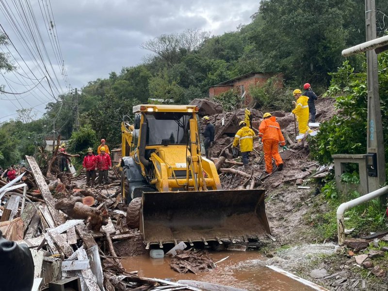 Fotografia colorida de um trator coletando destroços em um local alagado com trabalhadores de roupas amarelas e laranjas trabalhando em sua volta 