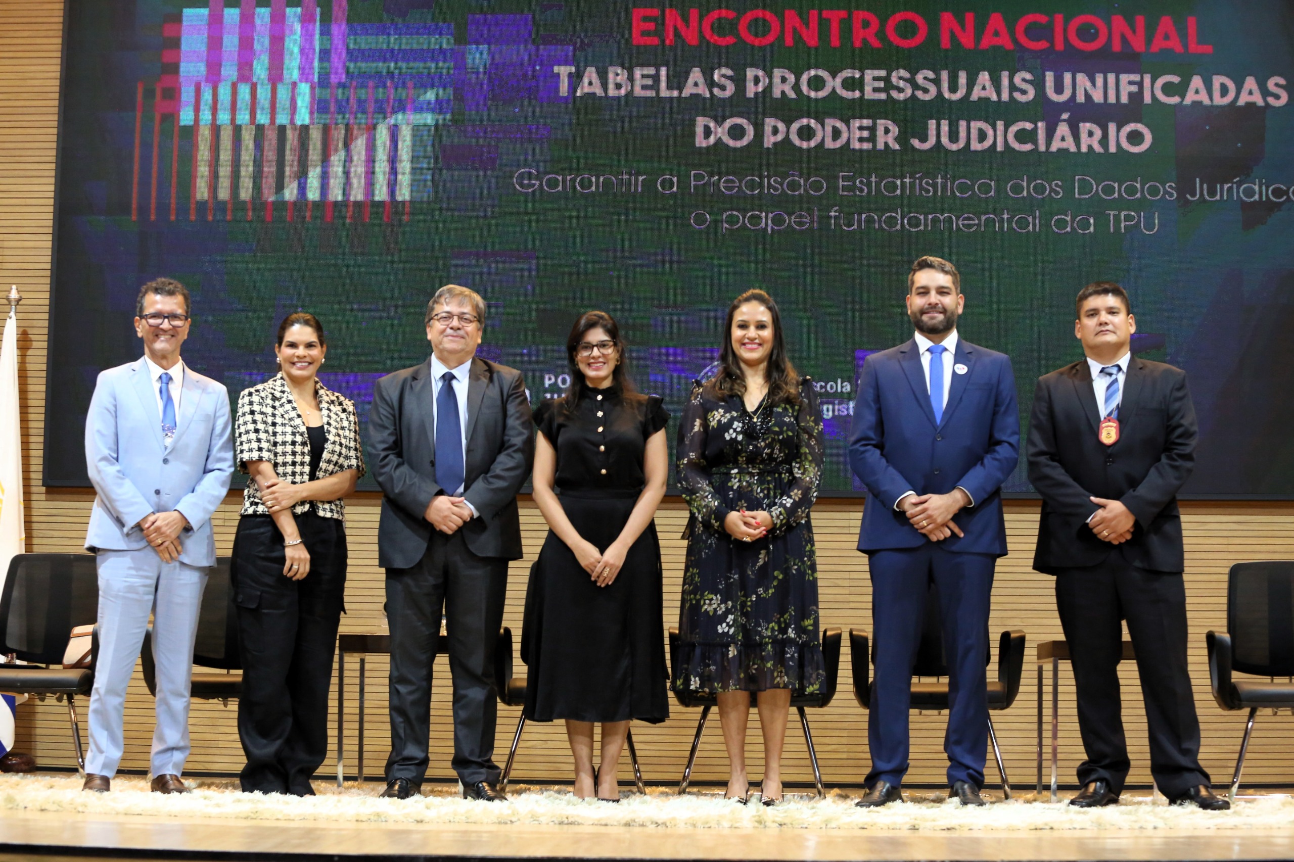 Fotografia colorida que mostra homens e mulheres em frente a um banner sobre Encontro Nacional de Tabelas Processuais Unificadas. Eles posam pra foto