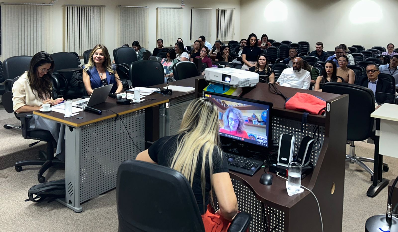 Imagem colorida que mostra três mulheres sentadas em uma sala de frente para uma plateia em um ambiente fechado