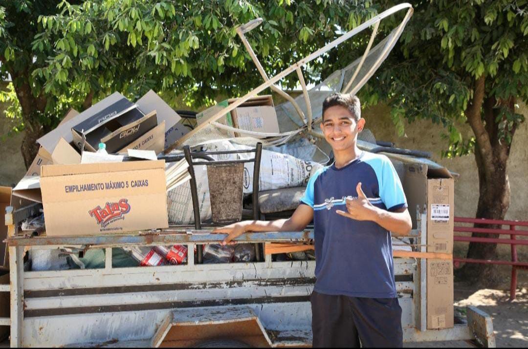 Fotografia colorida onde um menino de camisa azul está parado em frente a uma carretinha com alguns lixos recicláveis a algumas árvores ao fundo