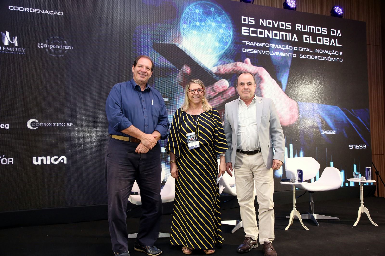 Fotografia colorida onde três pessoas, dois homens e uma mulher, posando para a foto de frente a um grande banner preto.