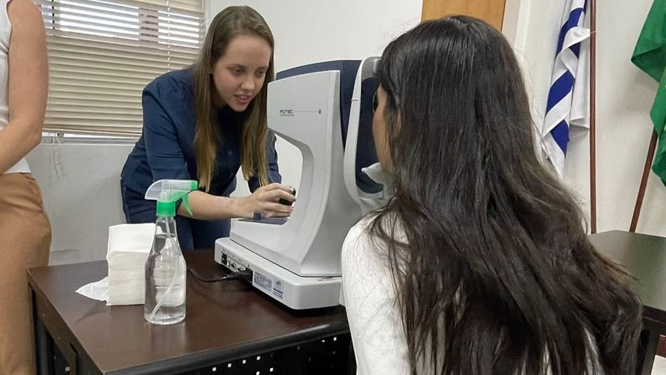 Mulher branca de cabelos castanhos claros e cumpridos, de blazer azul marinho faz exame de vista em aparelho em uma moça de cabelos lisos e longos, vestindo camisa de manga cumprida branca