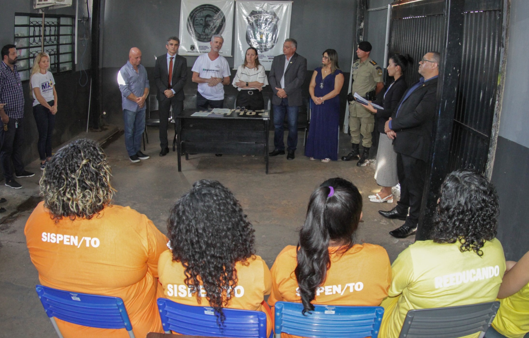 Fotografia colorida que mostra autoridades à frente de uma reunião e as reeducandas sentadas (se serem identificadas) ouvindo