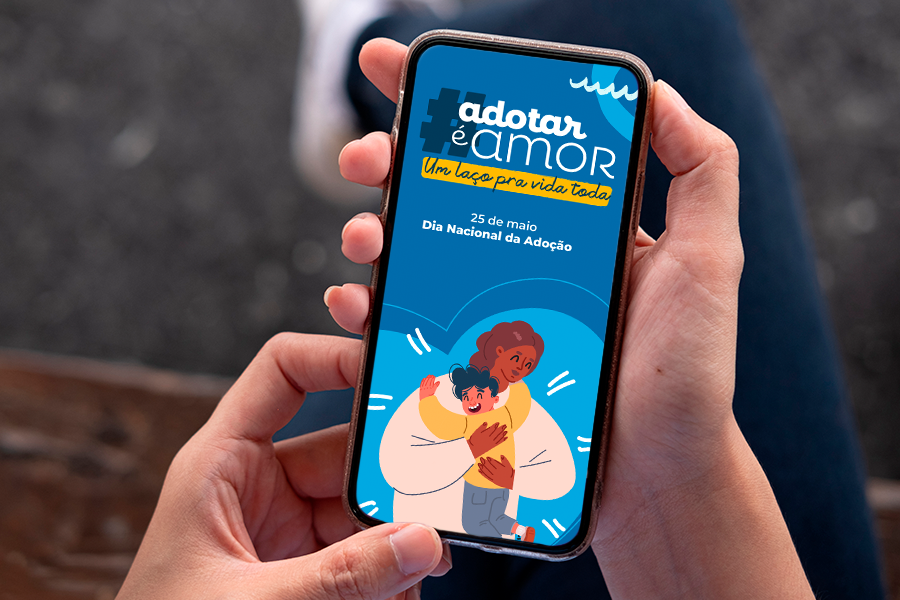 Na imagem aparece duas mãos segurando um aparelho celular e na tela  a figura de uma mãe com uma criança  com a hashtag adotar é amor um laço pra vida toda. Abaixo o texto 25 de maio Dia Nacional da Adoção