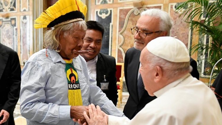 Cacique Raoni à esquerda com uma roupa branca e adereços indígenas na cabeça e no pescoço nas cores amarela, verde, vermelho, preto e branca, cumprimentando o papa (à direita com roupa branca) ao lado dois homens de terno escuro observam os dois