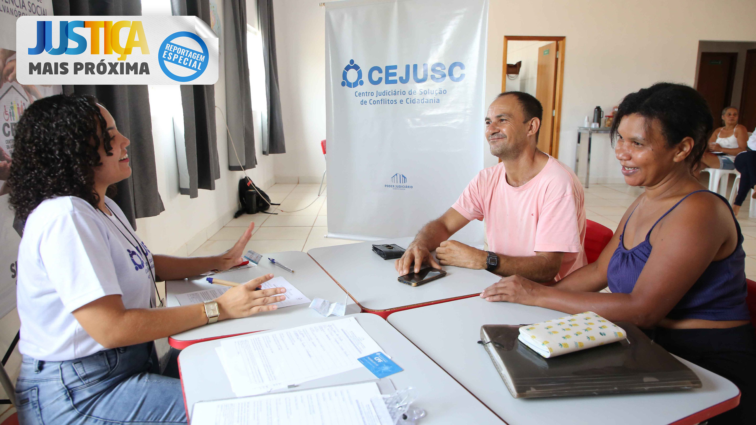 Em uma sala de atendimento, um casal, um homem moreno, de camiseta rosa, e uma mulher negra de blusa azul, é atendido por uma servidora; ao fundo um baner branco com o nome Cejusc