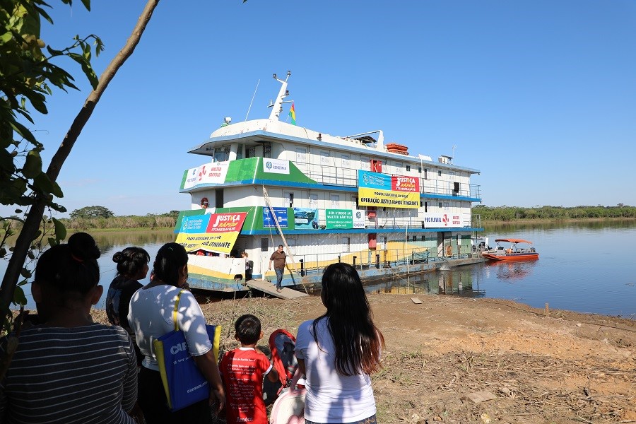 Imagem com um grande barco da justiça e um rio ao fundo, em primeiro plano pessoas de costa aguardando 