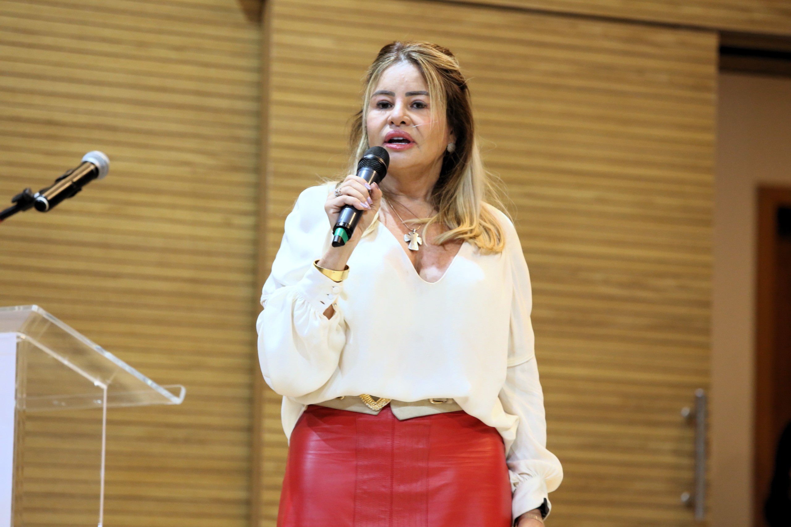 Juíza Milene de Carvalho, uma mulher de cabelos lisos e louros em pé, falando ao microfone; ela está de blusa branca e saia vermelha