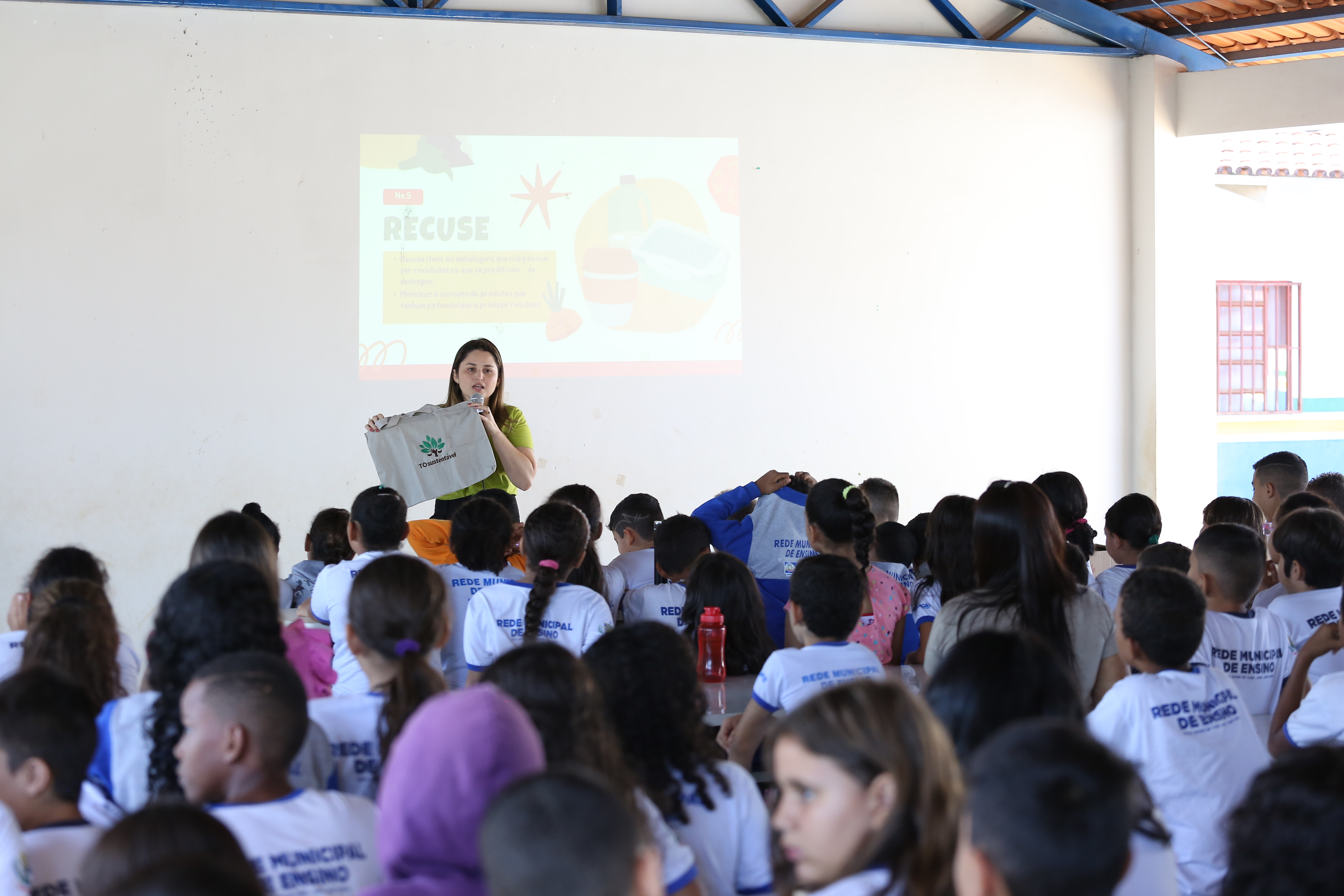 Mulher branca, cabelo liso, roupa verde e preta fala em uma sala cheia de alunos