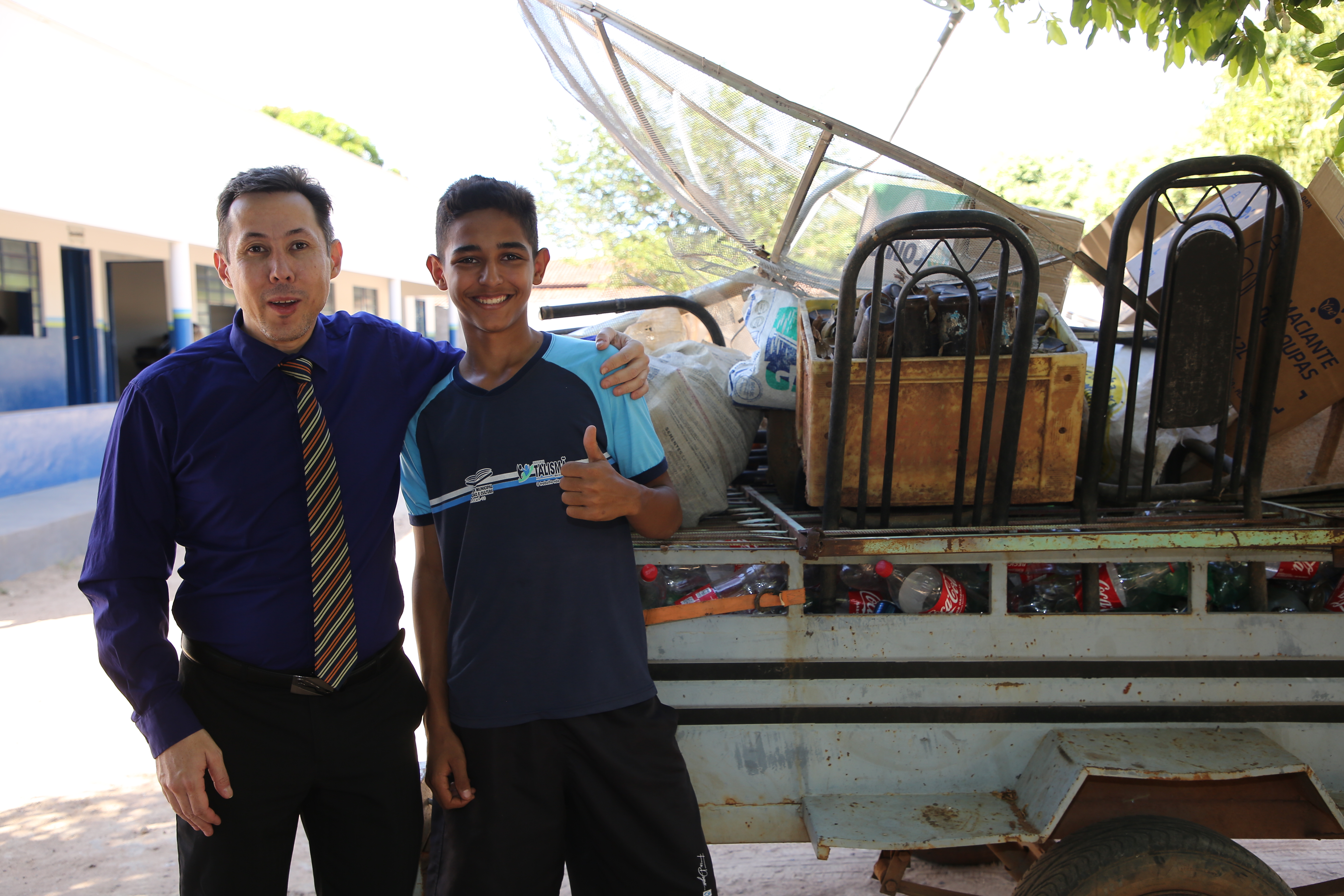 Imagem mostra um homem de roupa azul e preta e gravata e um jovem também com roupa azul e preta; atrás uma carreta com materiais recicláveis