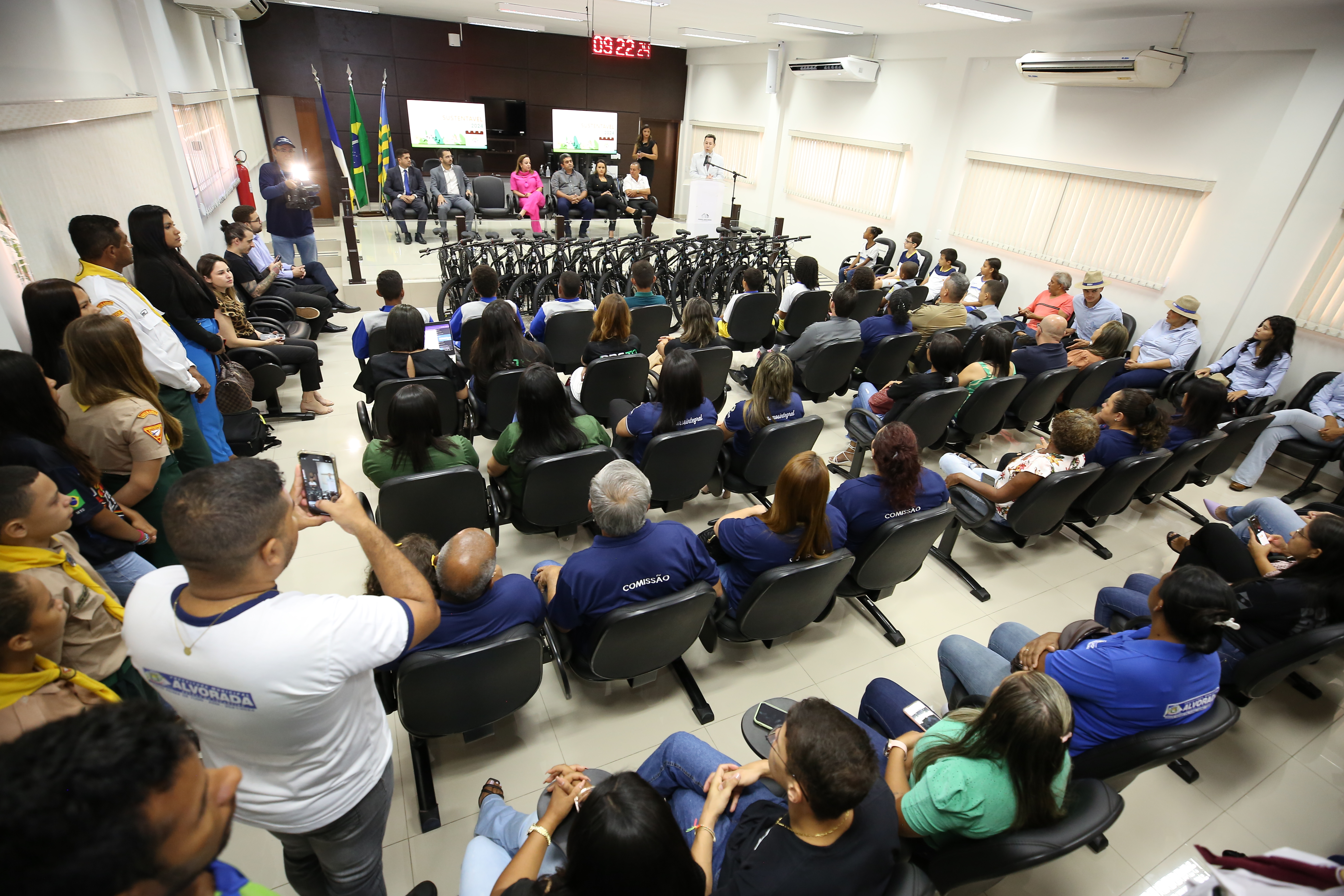Foto mostra um auditório com várias pessoas sentadas em cadeiras e à frente uma mulher de rosa discursa