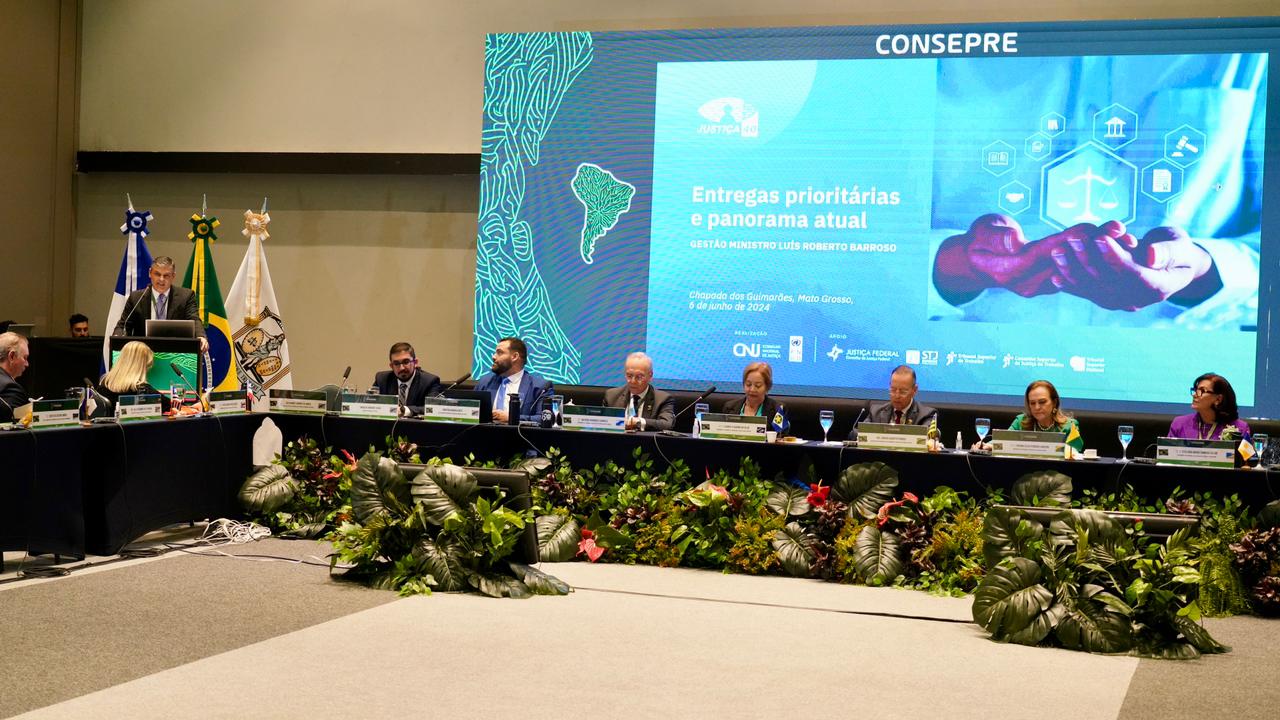 Homem de terno escuro fala ao púlpito no canto esquerdo, em frente a um mastro com 3 bandeiras. Há ainda uma mesa composta por desembargadores e desembargadoras do Brasil e ao fundo uma tela em LED fala do encontro destes desembargadores