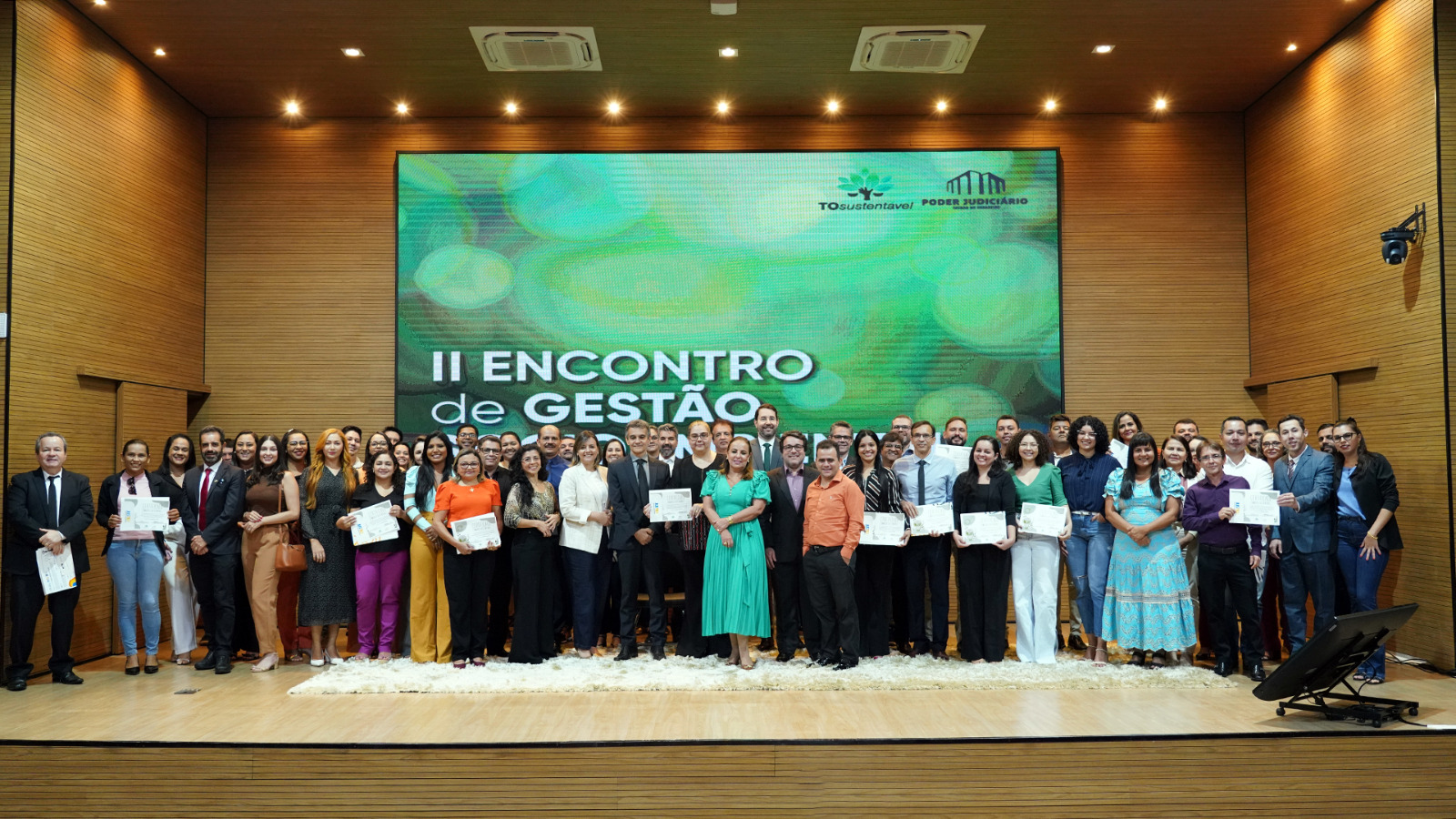 Fotografia colorida que mostra homens e mulheres em roupa social posando para fotografia segurando certificados. Eles estão dentro do auditório do TJTO e ao fundo tem um telão verde do qual lê-se: II Encontro de Gestão